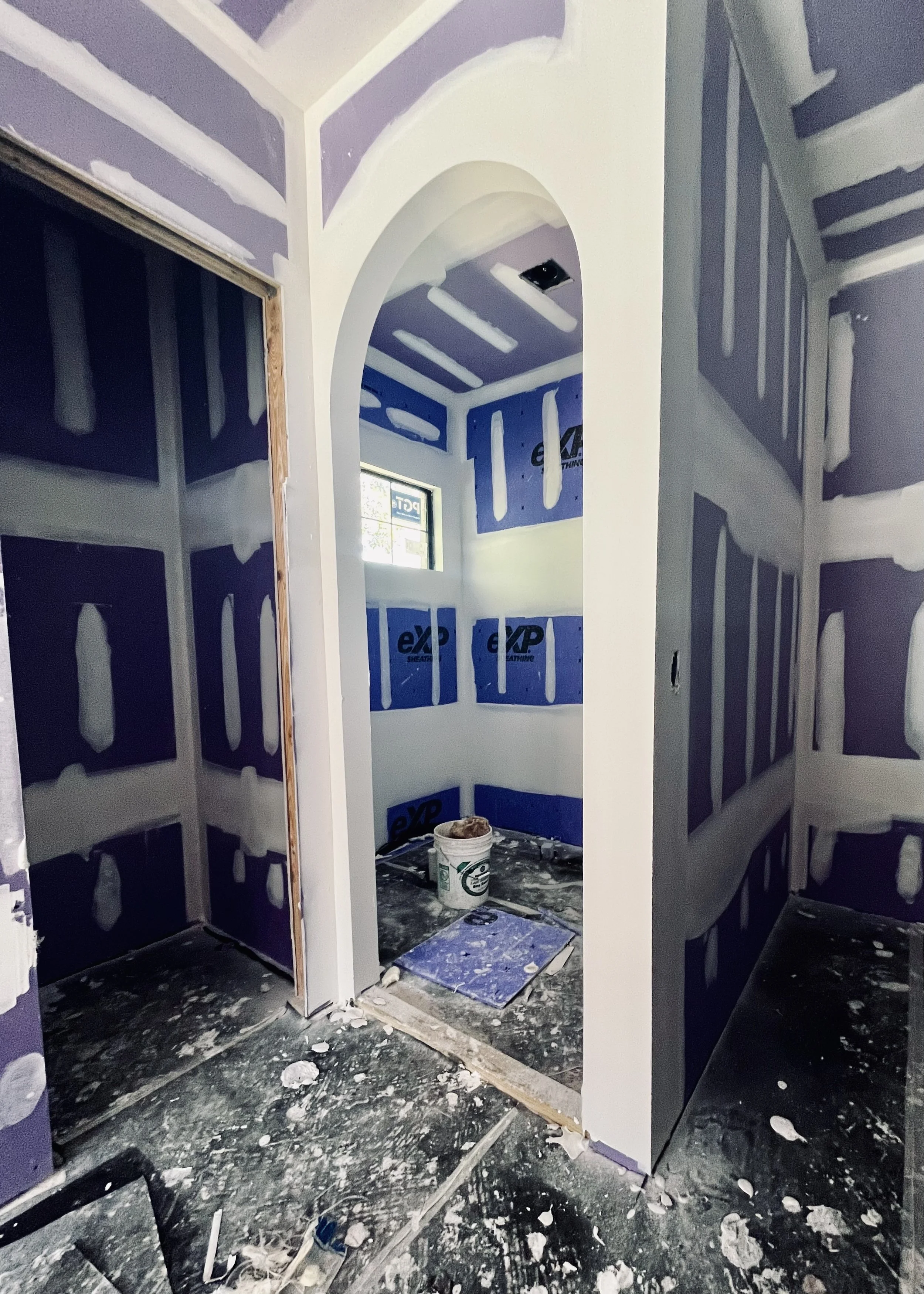 Interior of a building under construction with drywall installation, showing purple and white drywall, a partially finished arched doorway, a small window, and construction tools and materials on the floor.