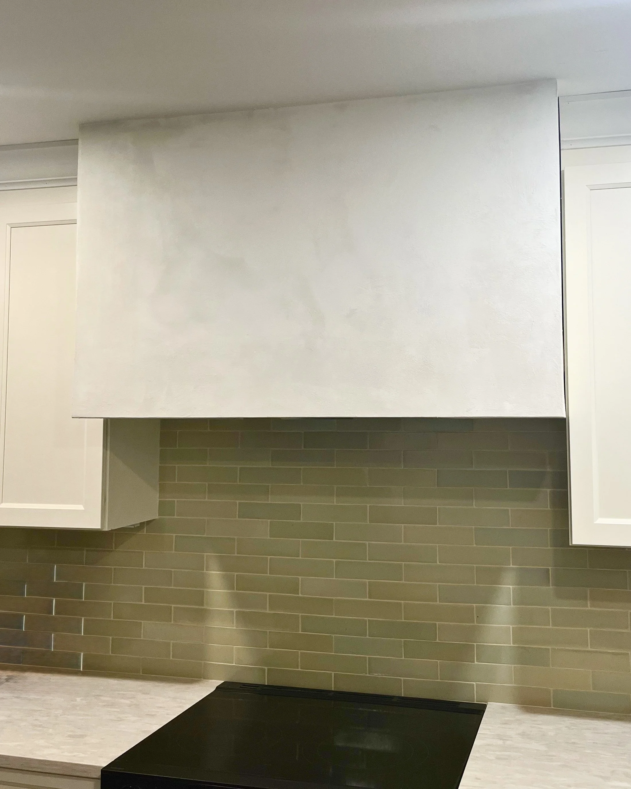 Kitchen with white upper cabinets, green tile backsplash, black stovetop, and white countertop.