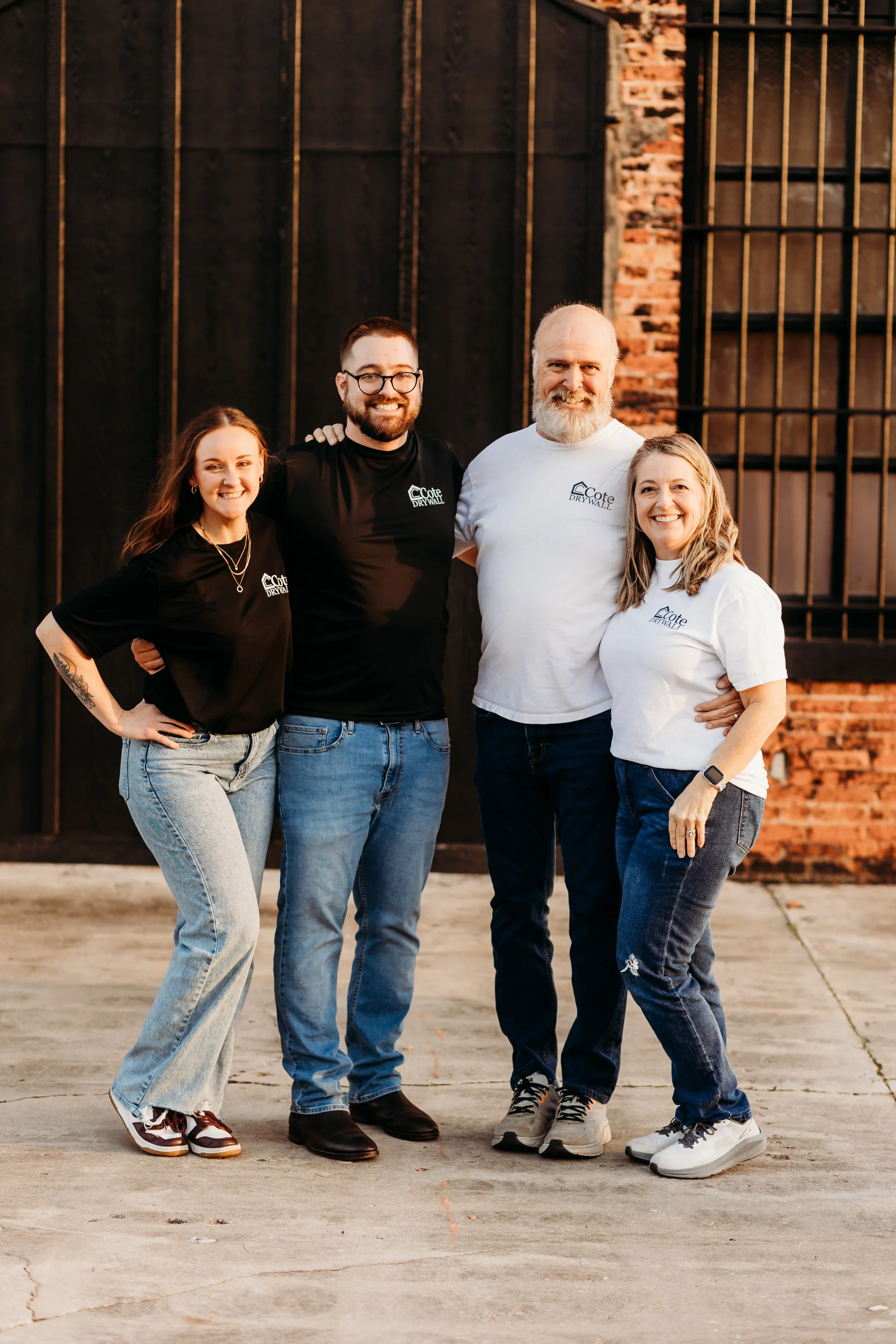 Four people standing together outdoors in front of a brick wall, smiling, wearing matching t-shirts with a company logo.