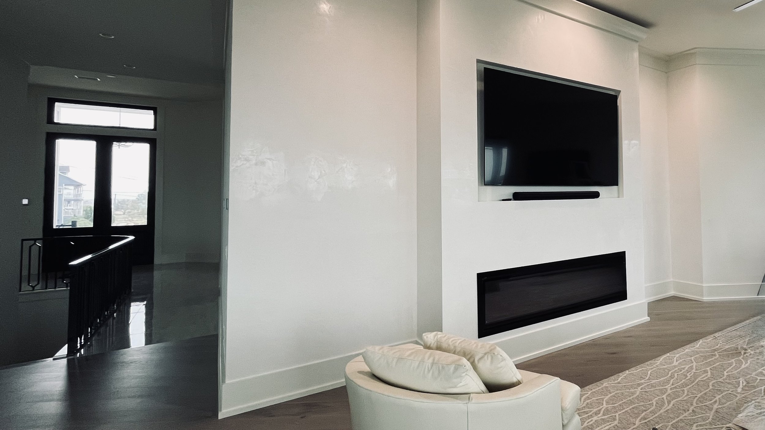 Modern living room with a wall-mounted flat-screen TV, a built-in fireplace below, a beige armchair with pillows, a patterned rug, and a view through a glass door to the outside.