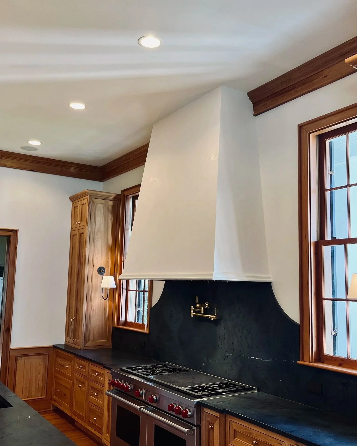Kitchen with wooden cabinets, black countertops, a range stove, large windows, and a sloped white vent hood above the stove.