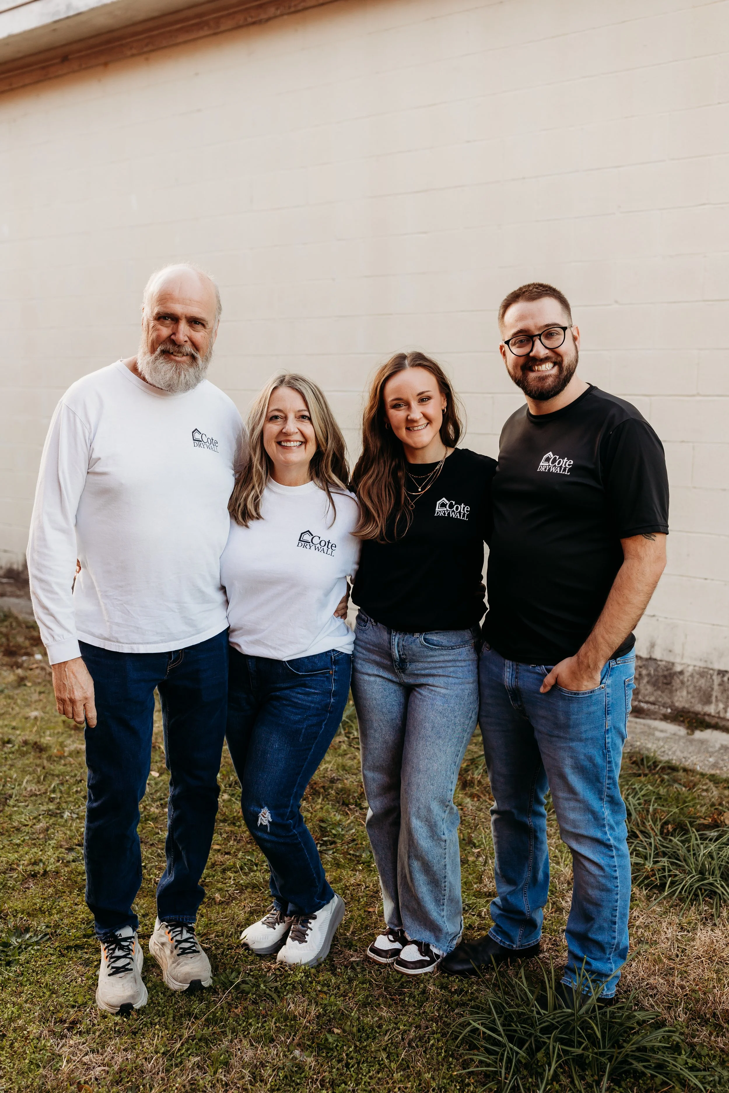 Group of four people standing outdoors in front of a beige wall, smiling for the camera, wearing matching 'Cote Drywall' shirts.