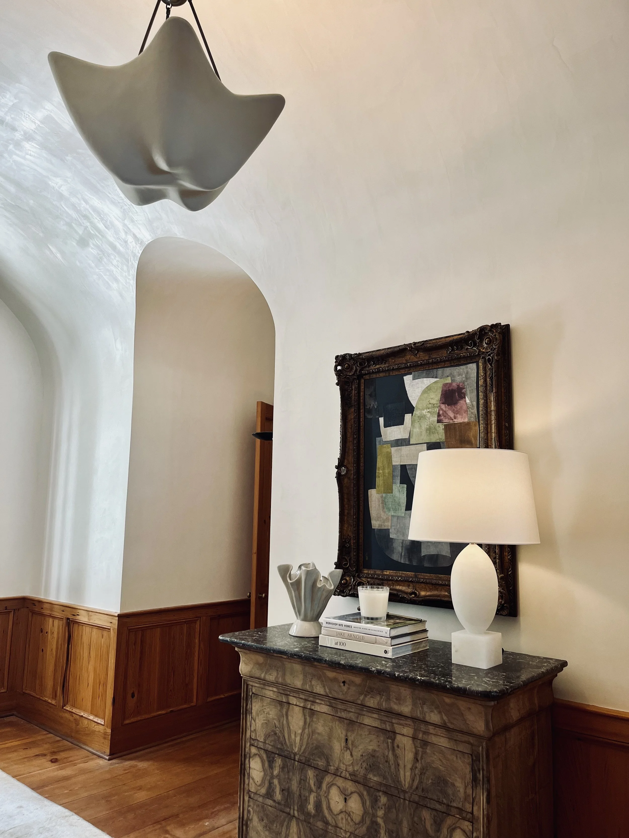 A living room corner with a wooden sideboard, a framed abstract artwork, a white table lamp, a stack of books, a vase, and a candle. There is a ceiling light fixture hanging from the ceiling.