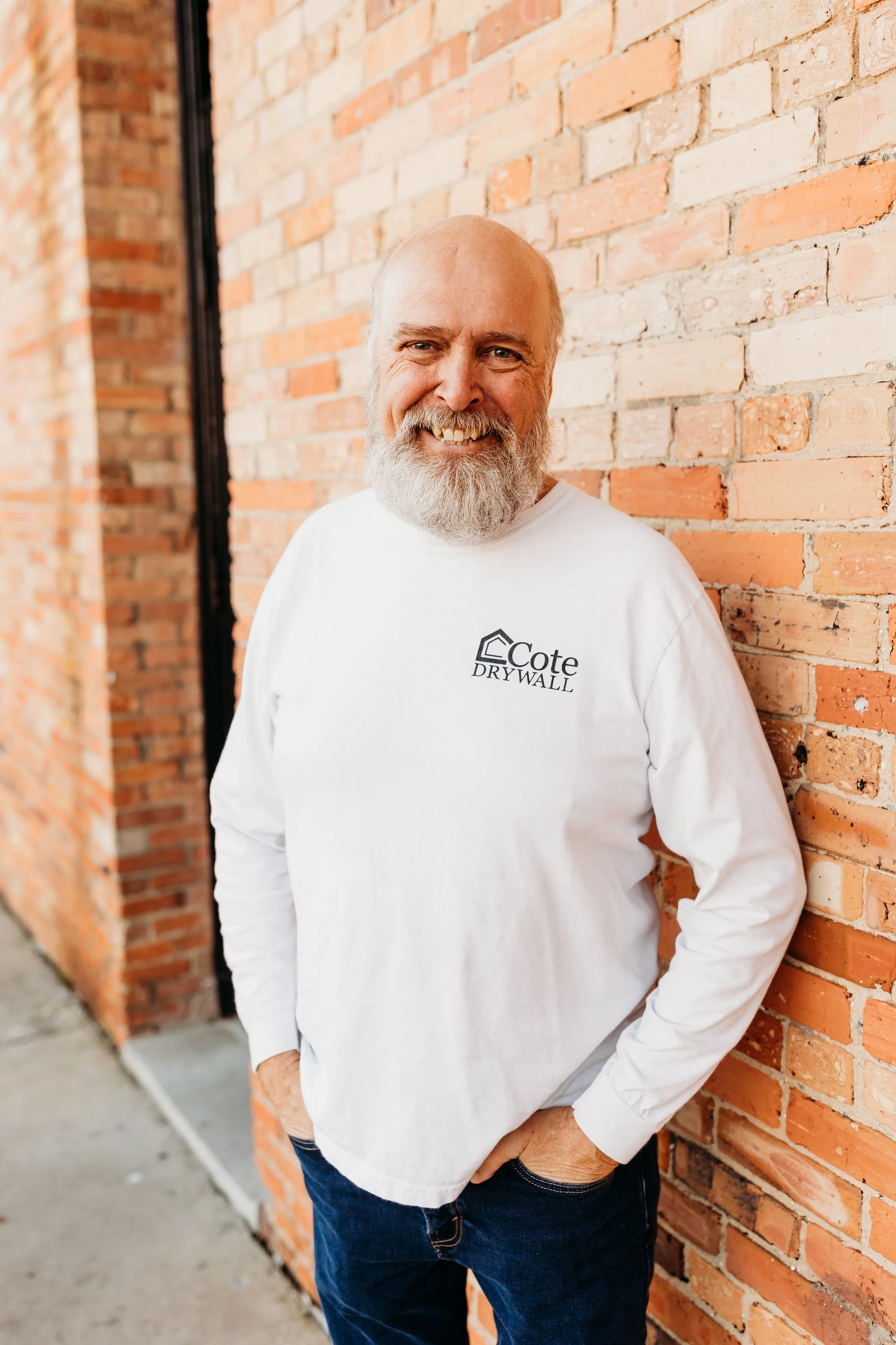 A smiling man with a beard, wearing a white long-sleeve shirt with a company logo, leaning against a brick wall.