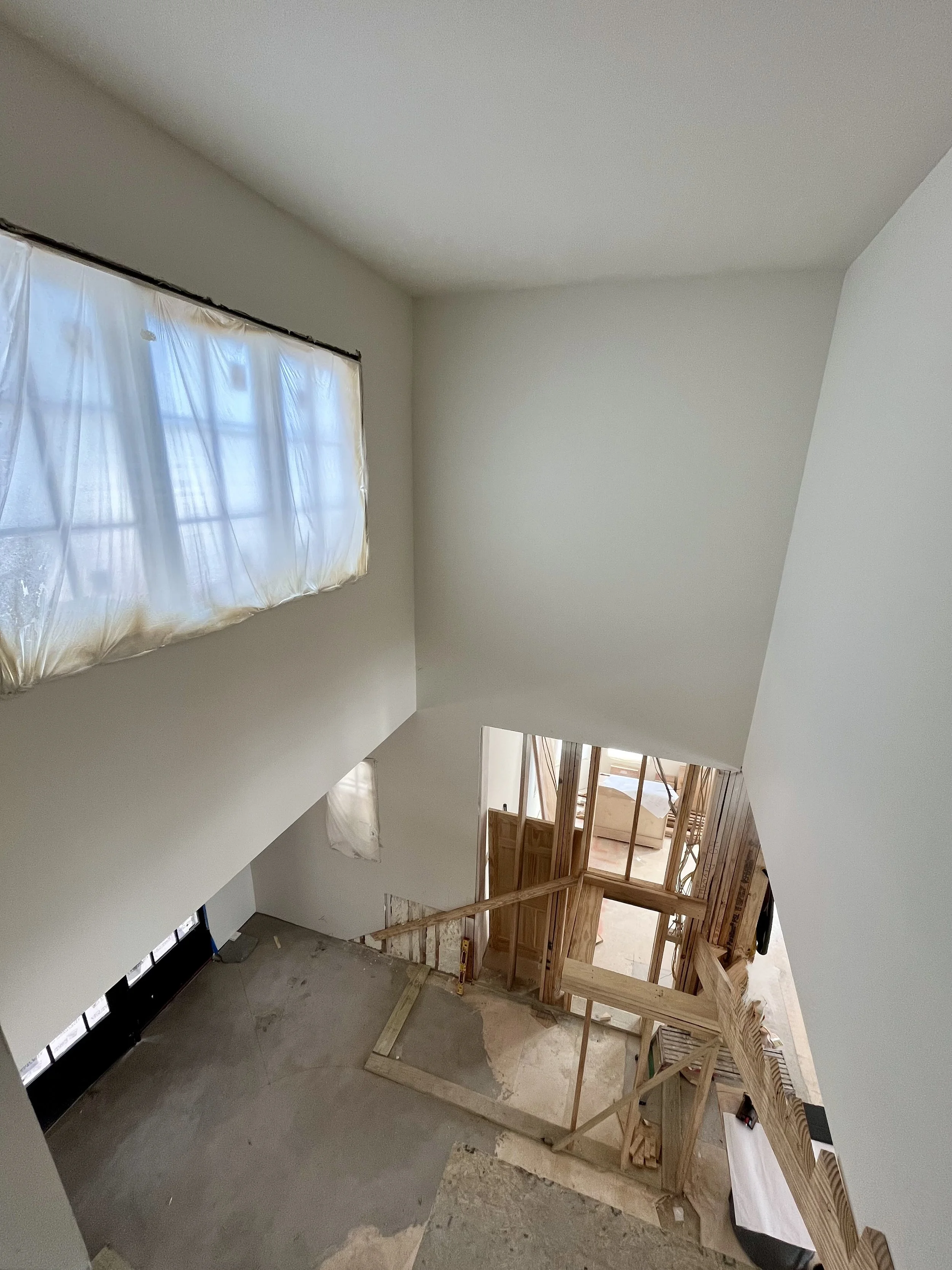 Interior of a home under construction, showing a staircase with unfinished wooden framing and an upper window with plastic covering, and an unfinished floor.