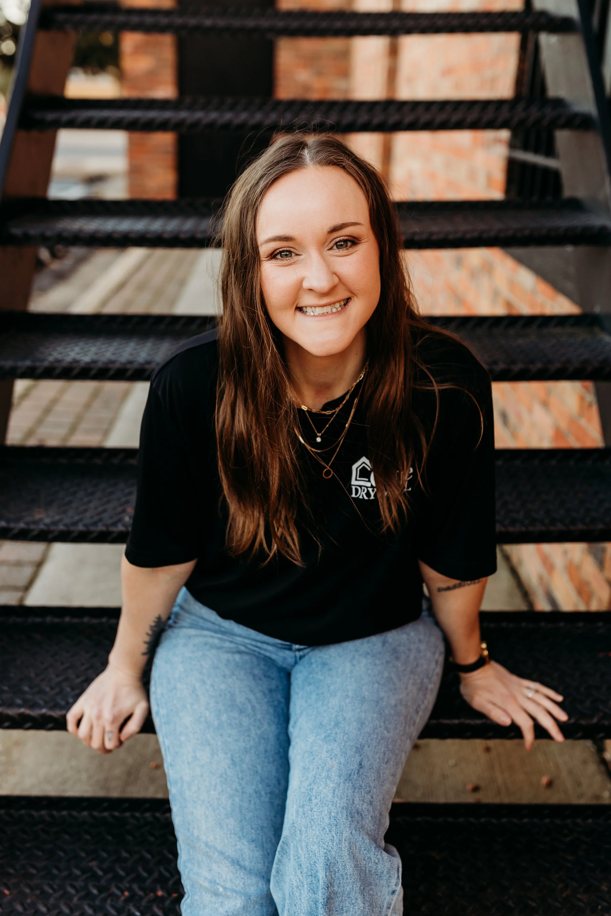 A young woman with long brown hair, smiling, sitting on black metal outdoor stairs, wearing a black T-shirt and light blue jeans.