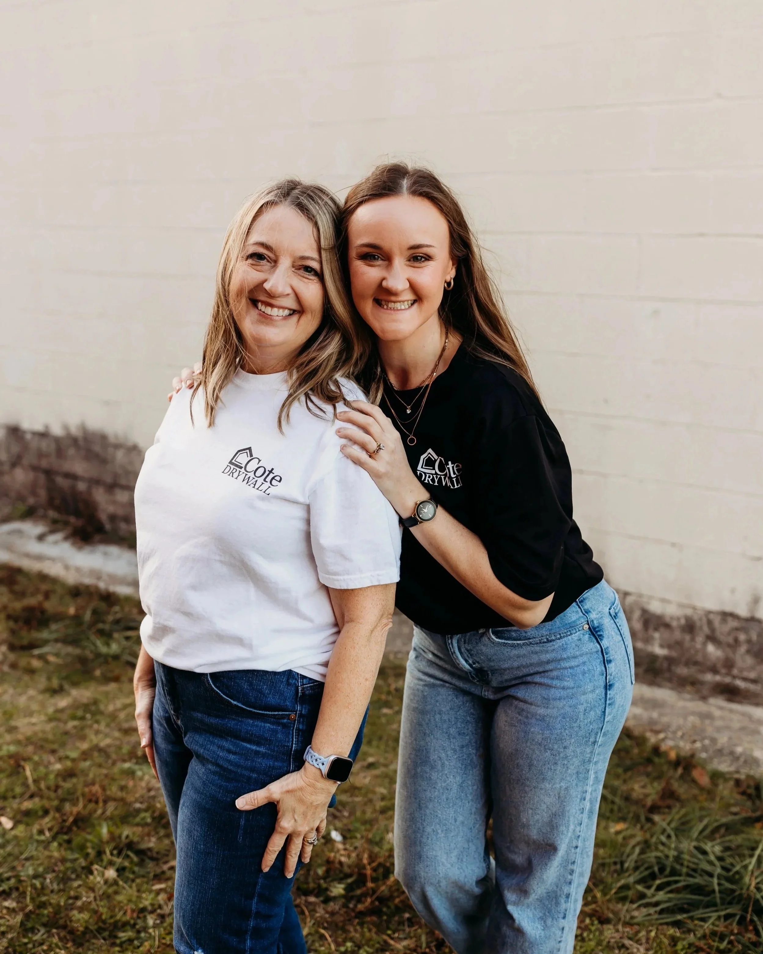 Two women smiling and posing outdoors, one with a white T-shirt and the other with a black T-shirt, both wearing jeans and standing in front of a beige wall.