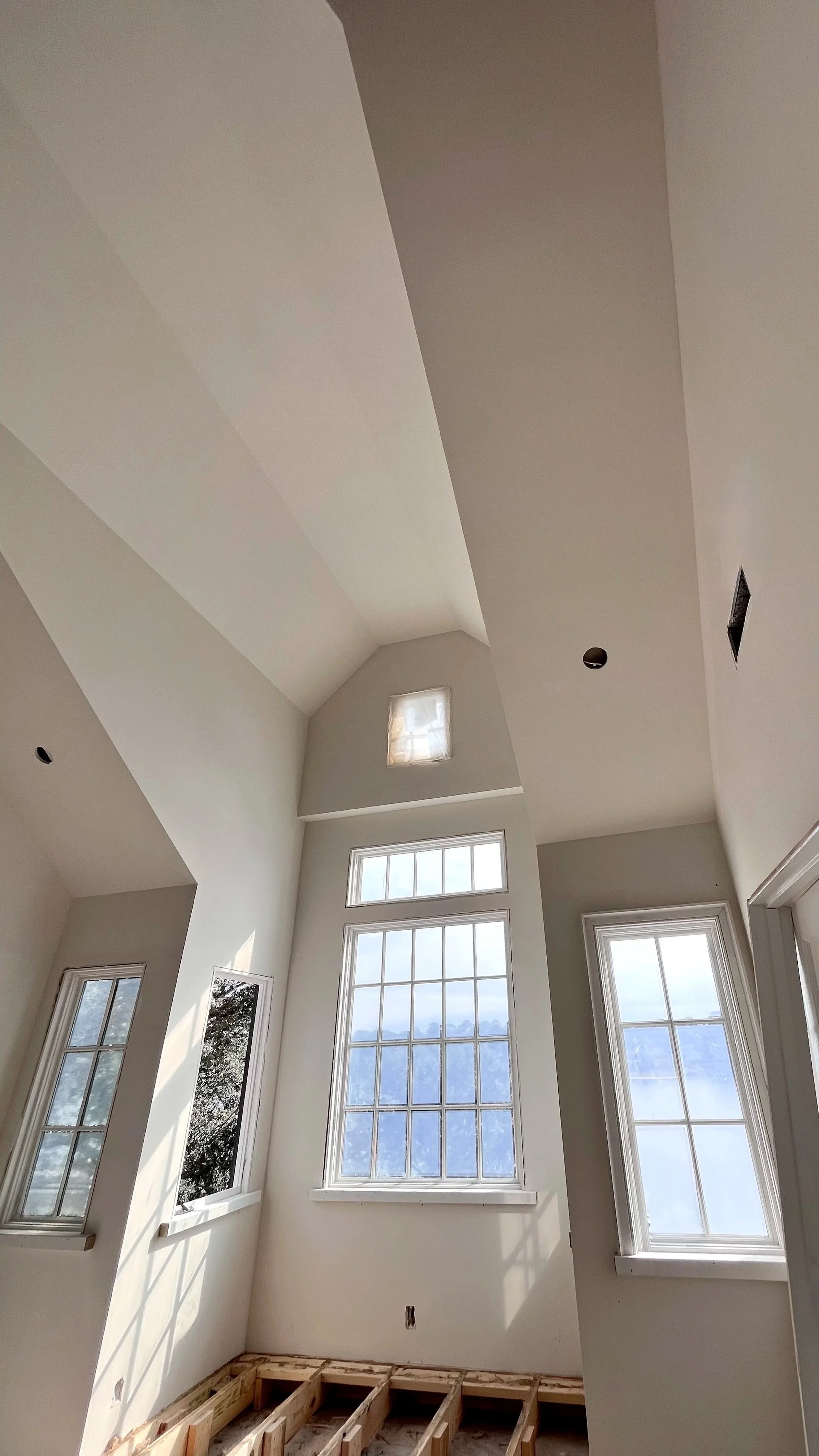 Interior of a room under construction with high ceilings, large windows, and exposed wooden floor framing.