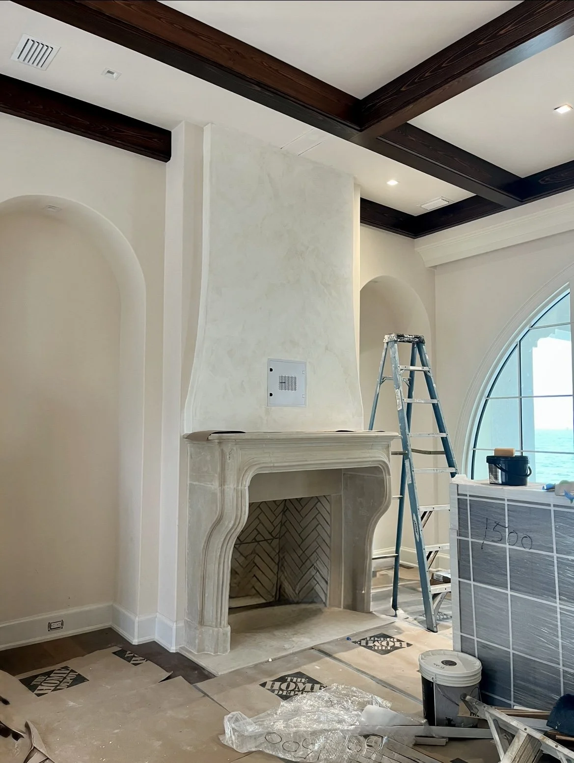 Interior of a room under renovation with a fireplace, stepladder, painter's tape, and construction materials, near a large arched window with ocean view.