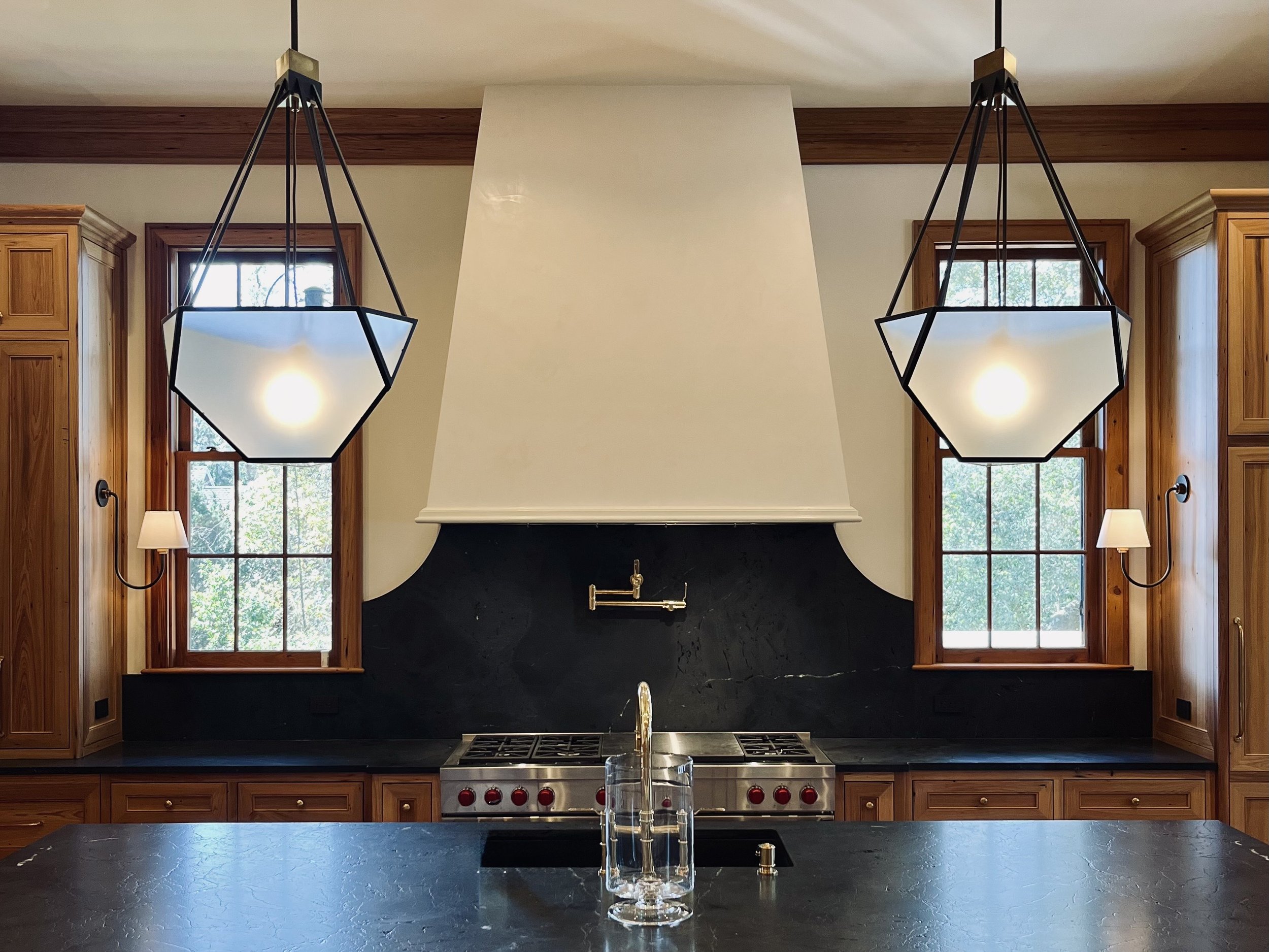 Kitchen with black countertop, wooden cabinets, two large windows, modern pendant lights, black and white range hood, gas stove, and a faucet with a glass.
