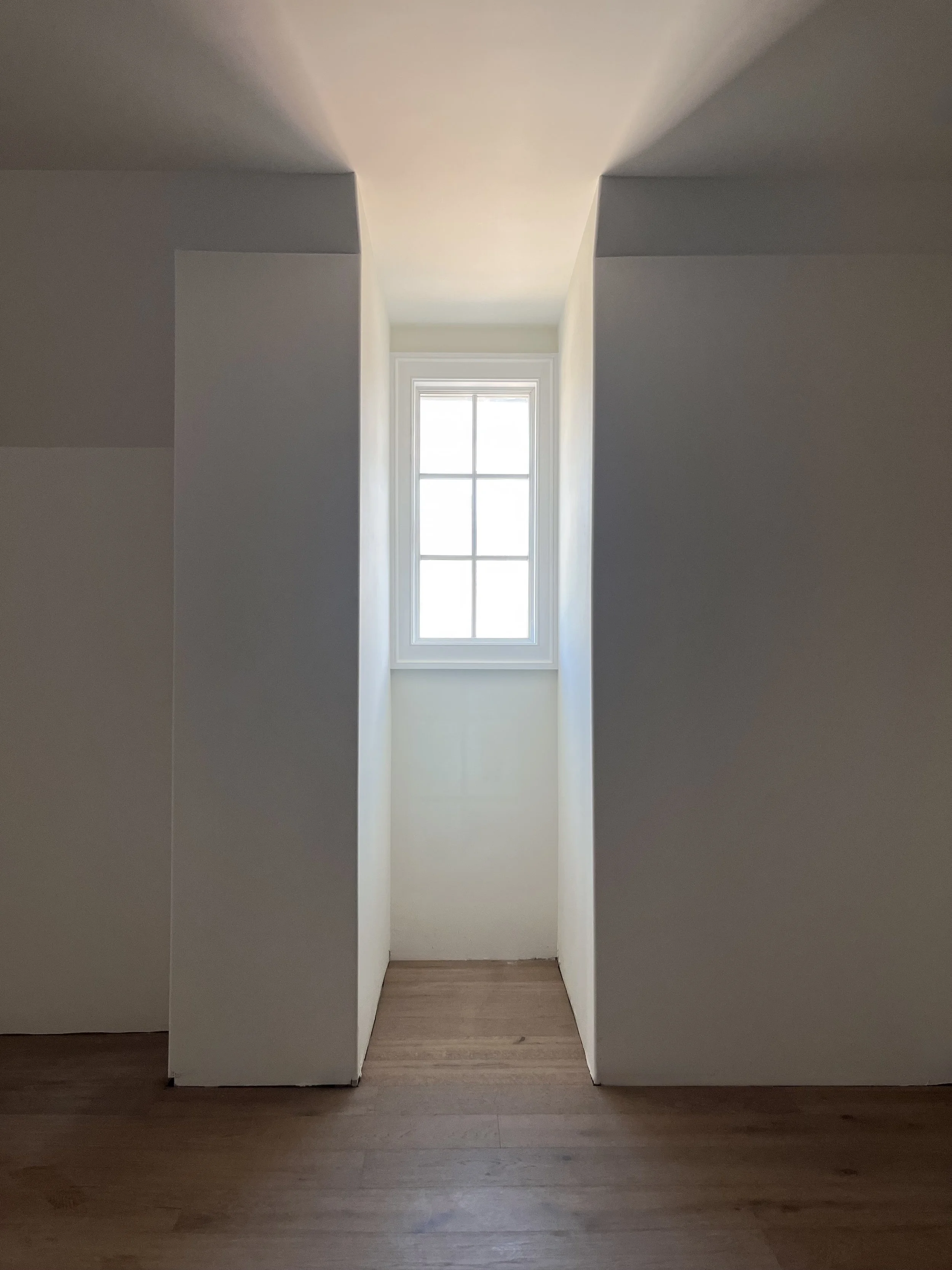 A narrow alcove with a single small window at the back, framed in white, in a room with white walls and wooden flooring.