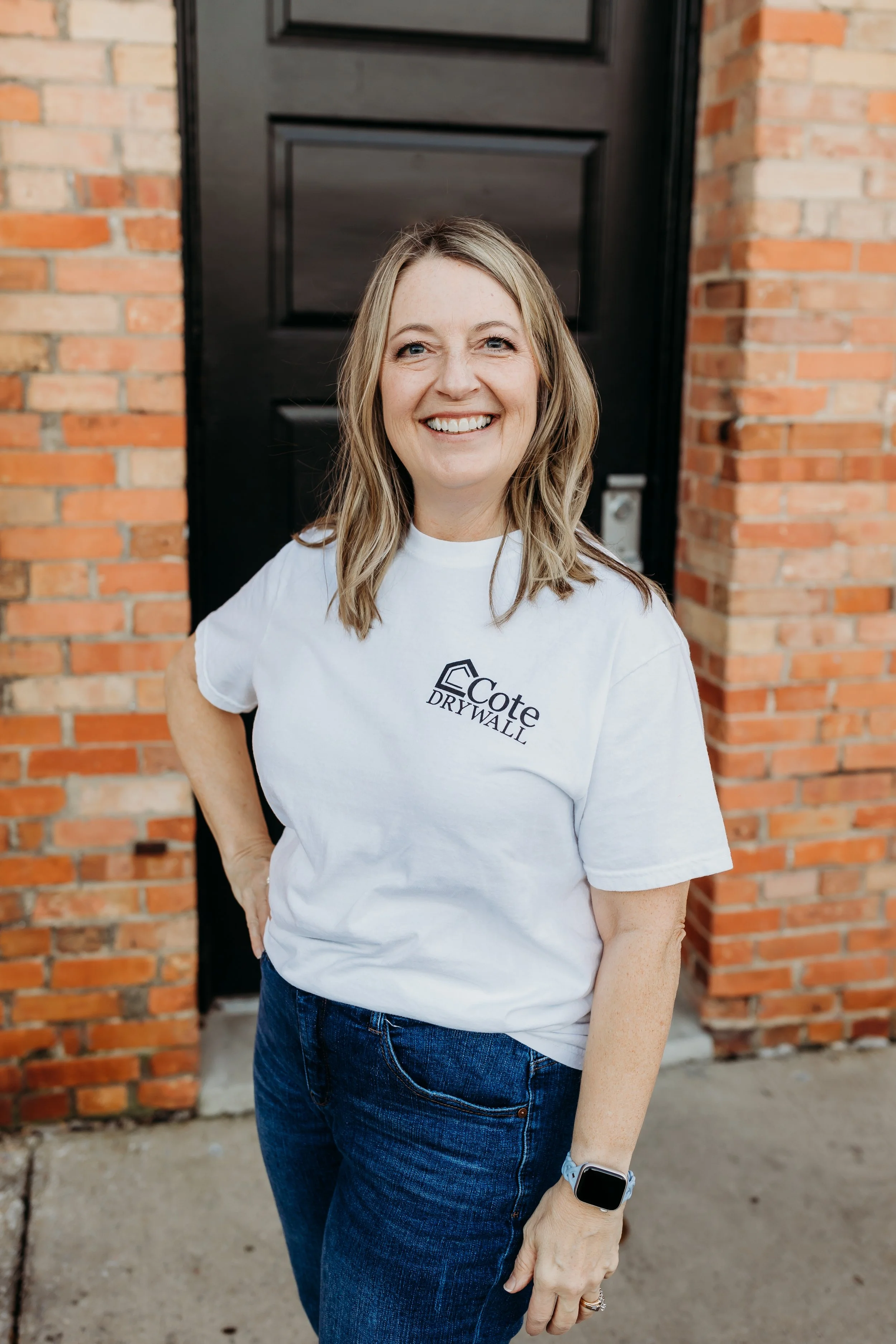 A woman with light brown, wavy hair smiling in front of a brick wall and a black door, wearing a white t-shirt with a logo and text that reads 'Cote Drywall', blue jeans, and a smartwatch.
