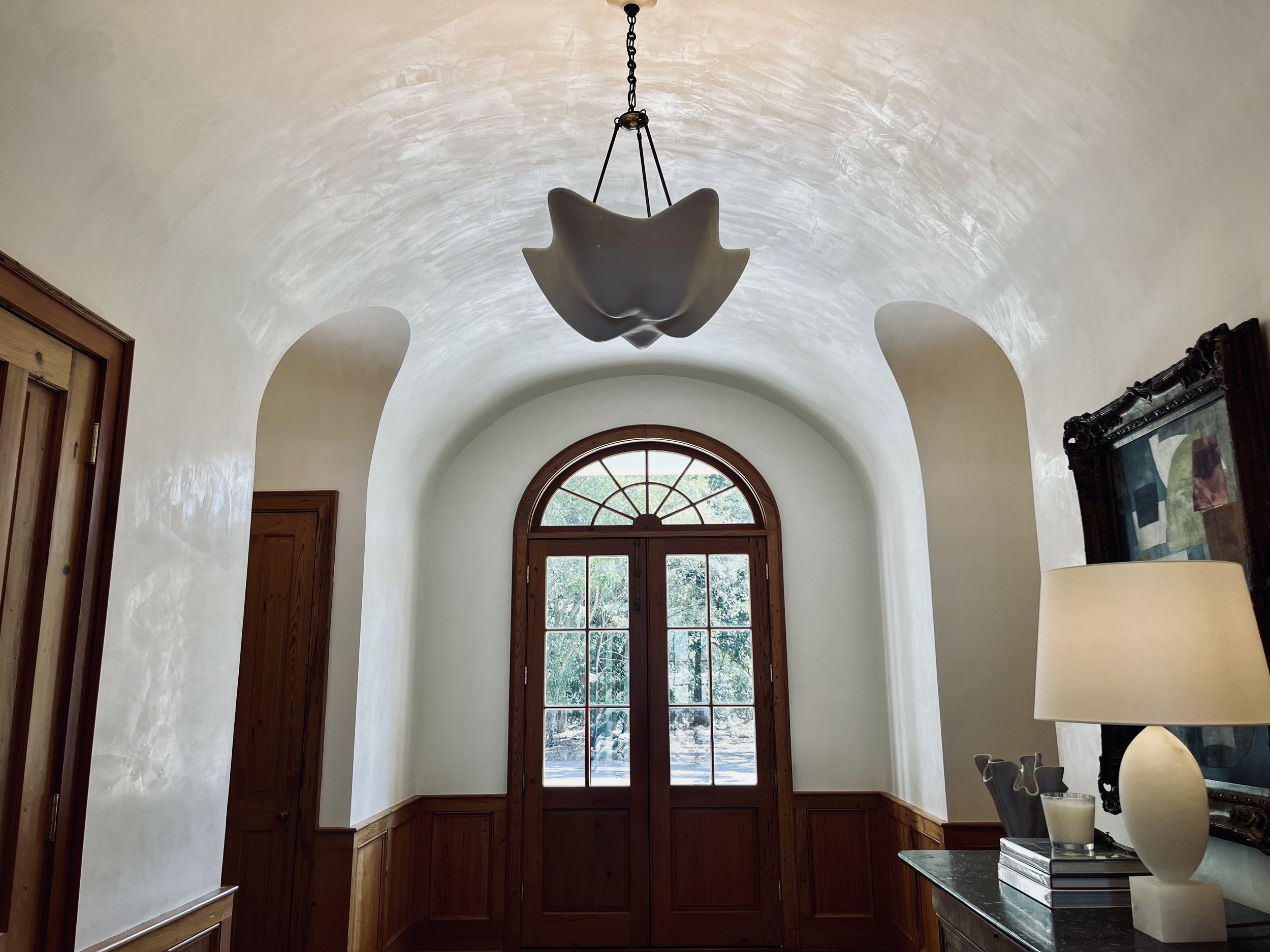 Interior view of a hallway with arched walls, wooden trim, a large window with an arched top, a modern pendant light fixture hanging from the ceiling, a lamp on a dark-colored console table, and framed artwork on the wall.
