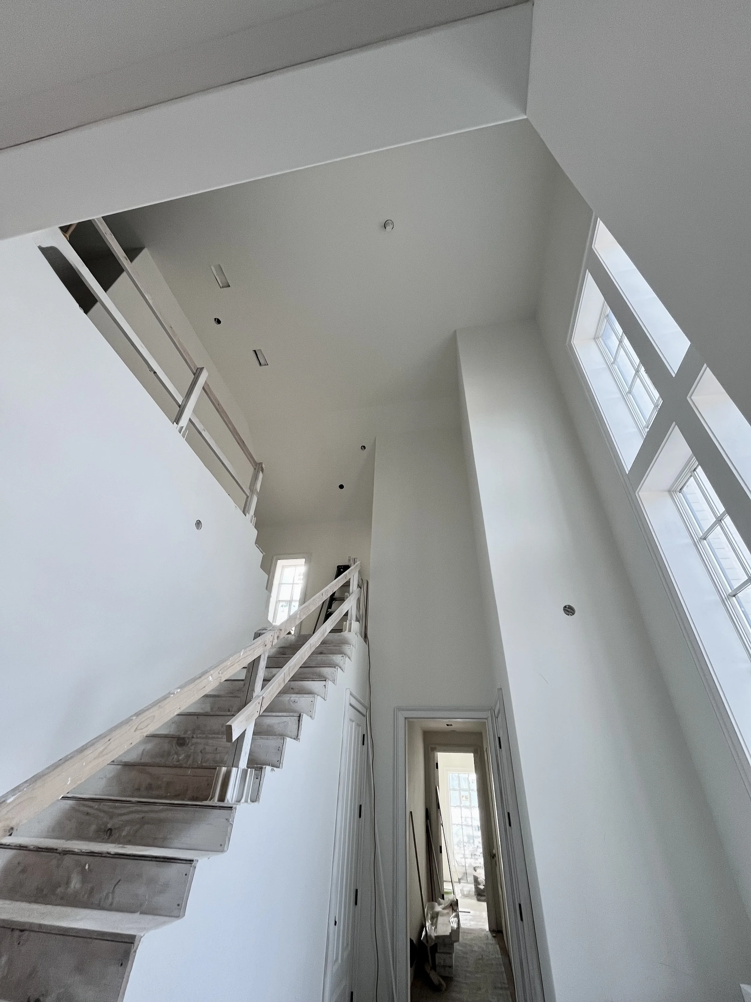 Interior view of a new house under construction, showing a staircase with unfinished steps and a high ceiling with large windows on the right, allowing natural light to fill the space.