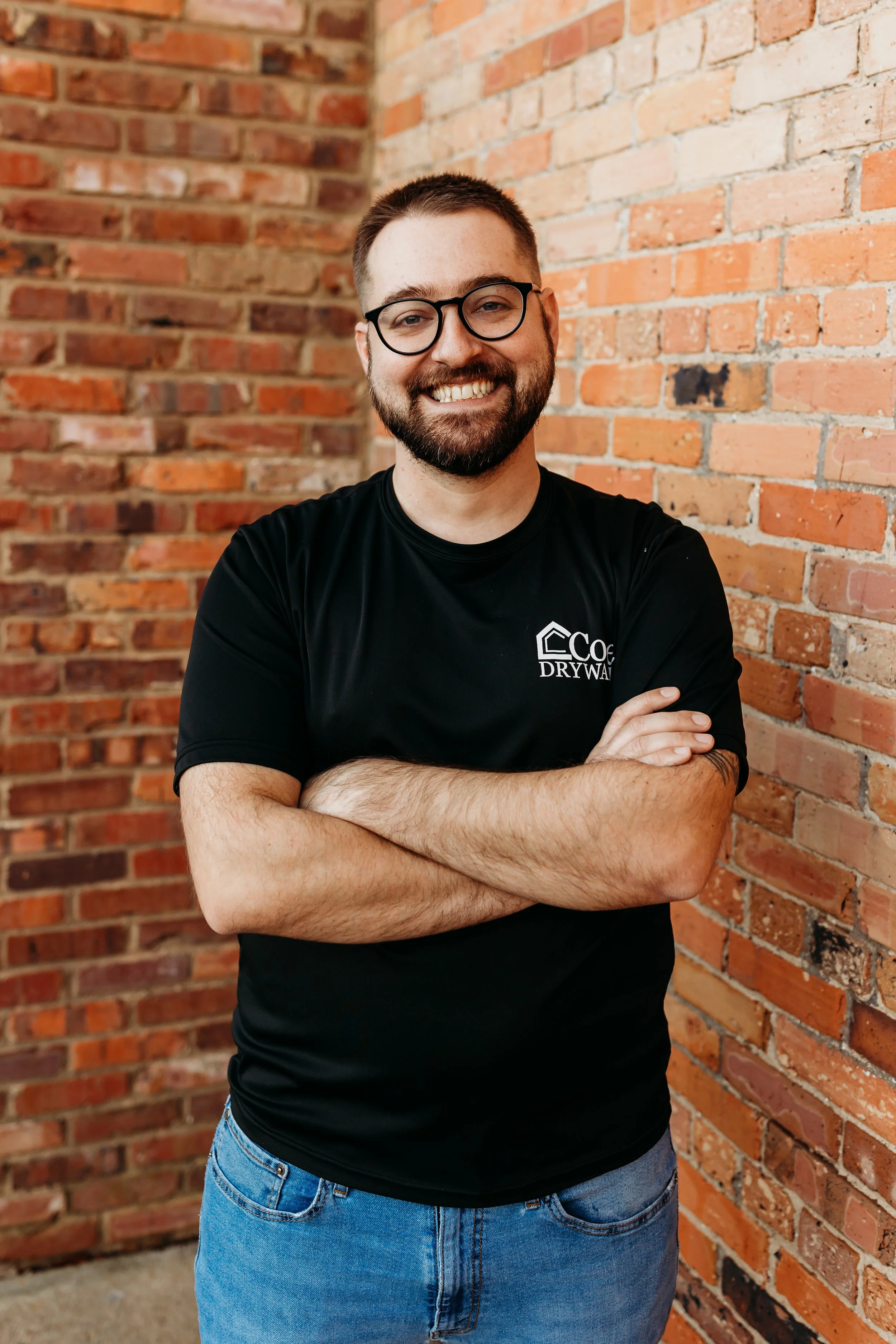 A smiling man with glasses and a beard, wearing a black T-shirt with a logo and crossed arms, standing in front of a brick wall.