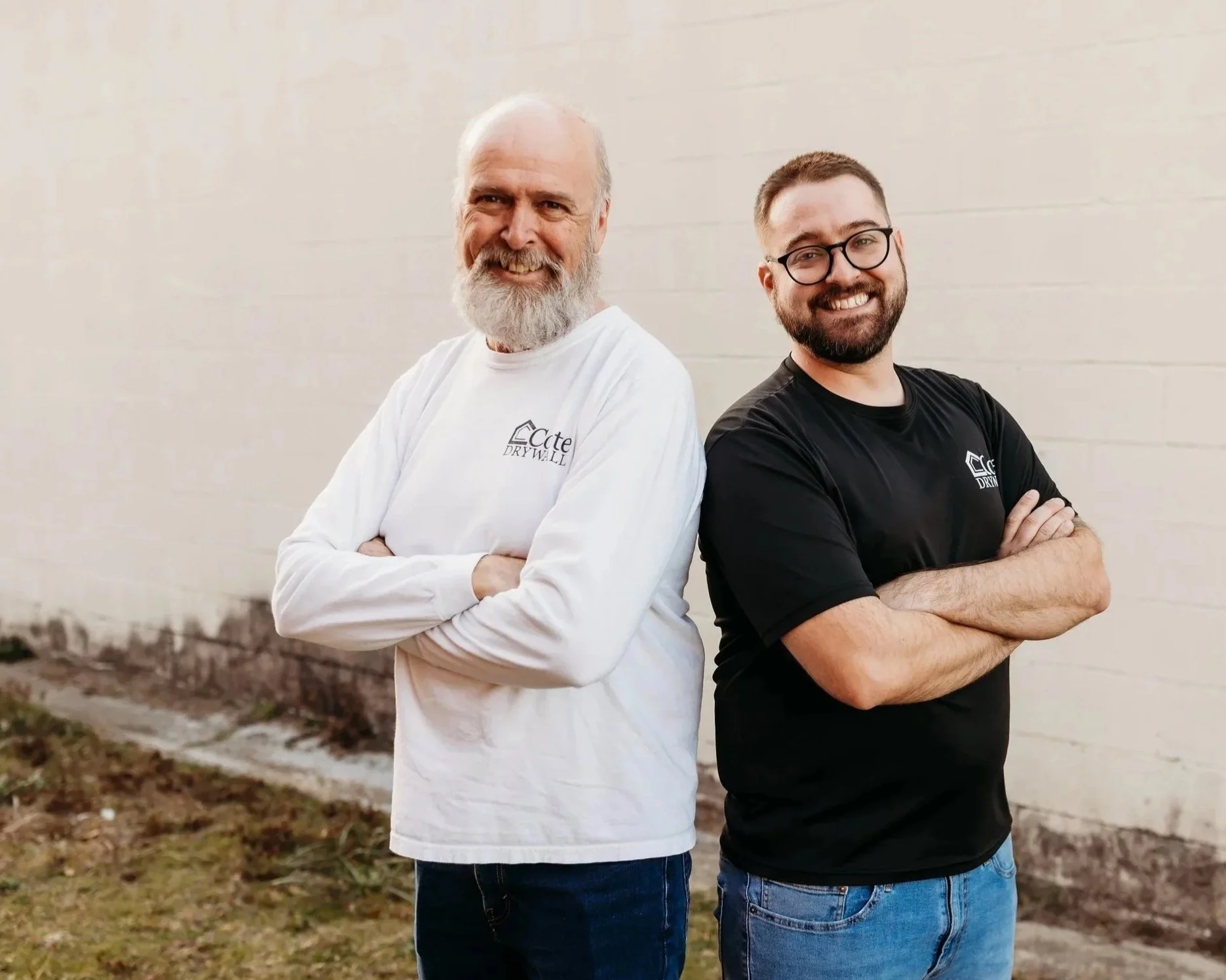 Two men standing outdoors against a beige wall, smiling with arms crossed, one with a beard and glasses, wearing a black T-shirt, and the other with a white beard, wearing a white long-sleeve shirt.