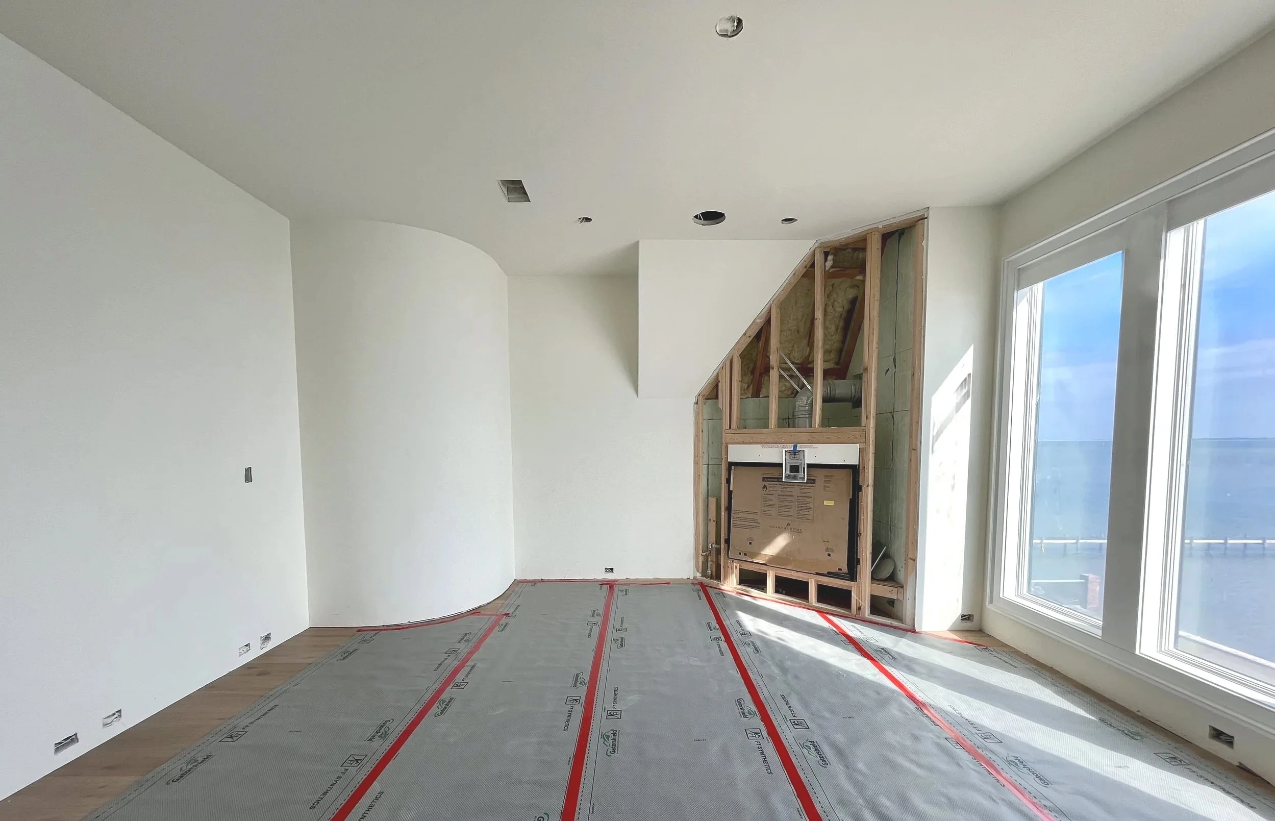 Interior of a room under construction with white walls, large windows on the right, and a partially built wall or fireplace at the far corner, with construction materials and insulation visible.