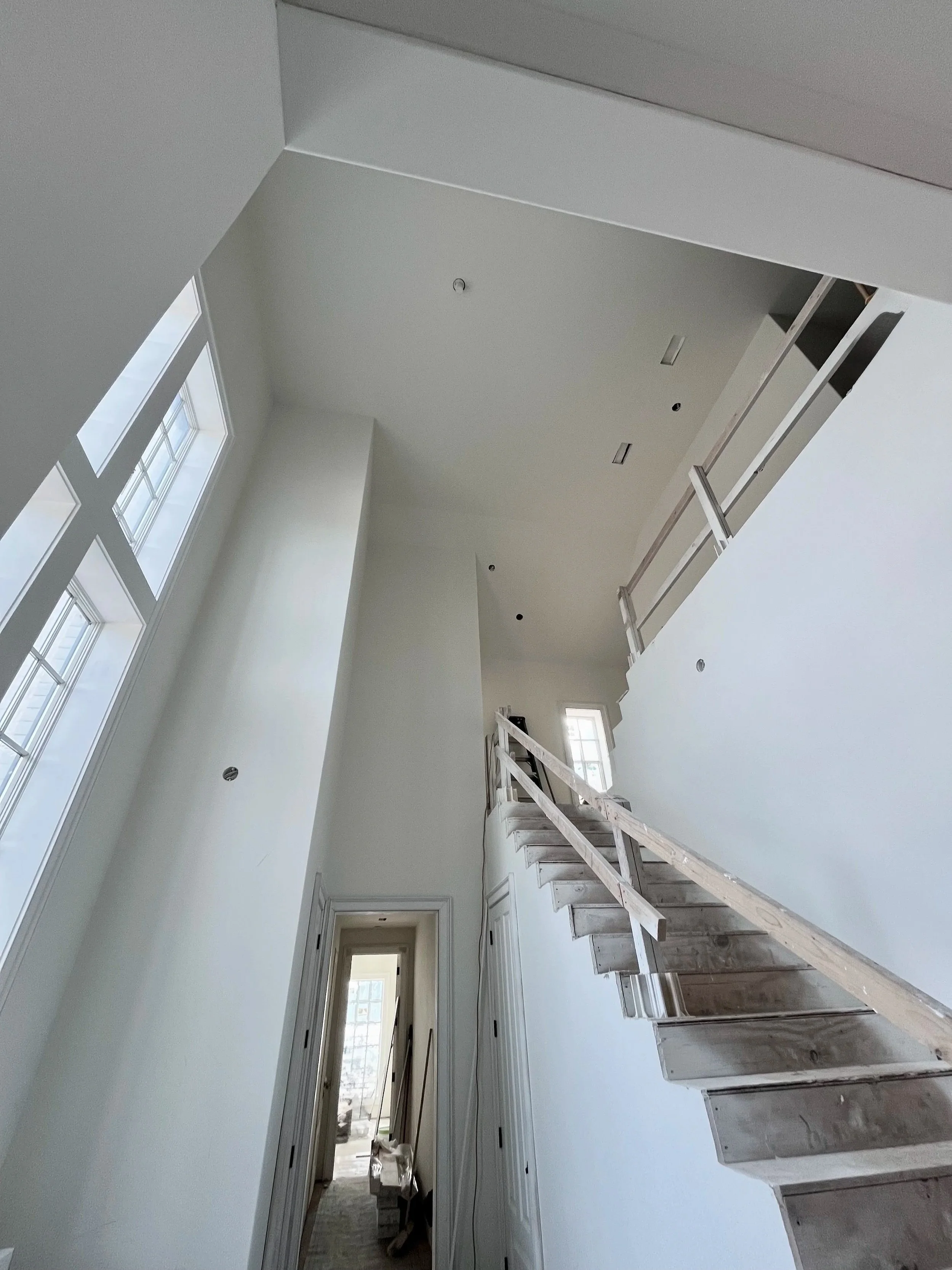 Interior view of a high-ceiling house under construction, showing a staircase with unfinished wooden steps, tall windows letting in natural light, and white walls.