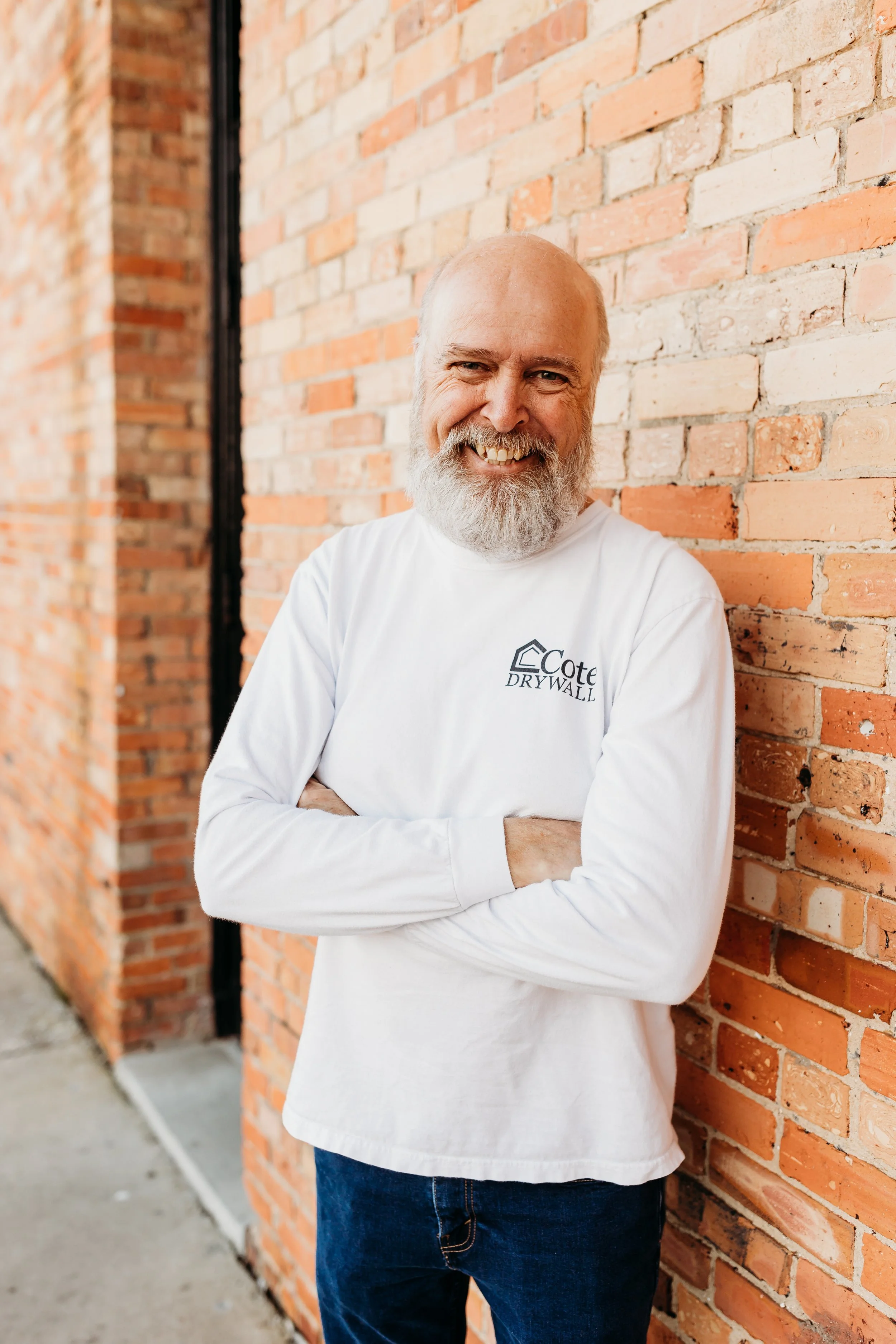 A smiling elderly man with a gray beard and bald head stands with arms crossed against a brick wall. He wears a white long-sleeve shirt with the logo 'Cote Drywall' on it.