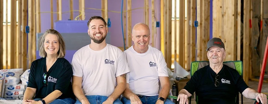 Four people sitting in front of a construction site, smiling, with exposed wooden framing behind them.
