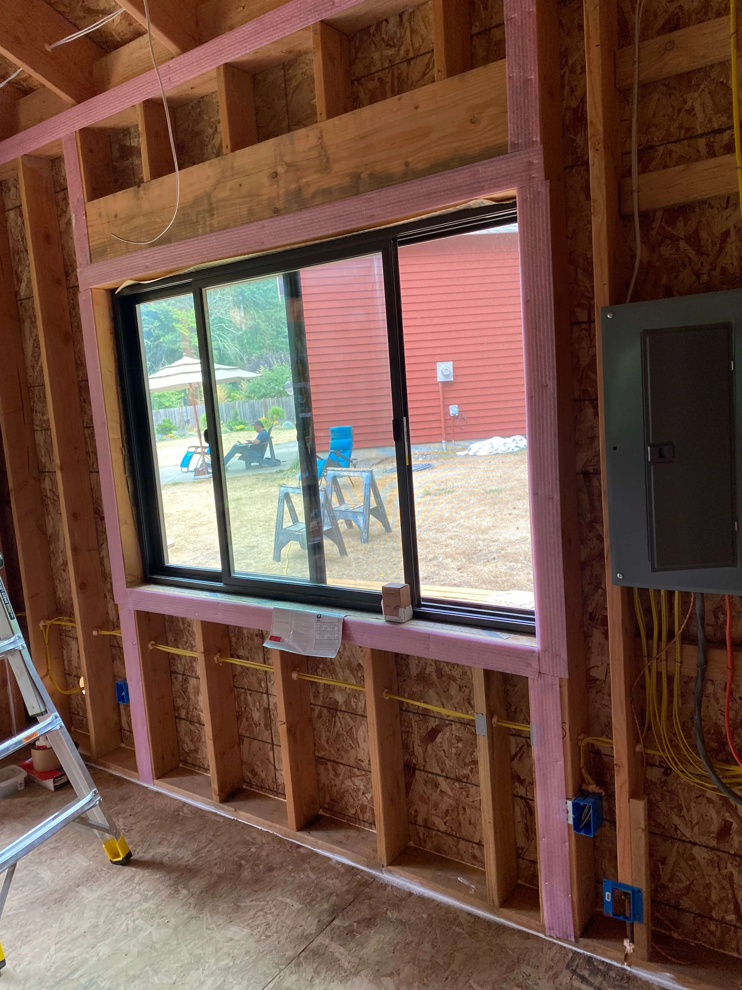 Interior view of a house under construction showing a large window with pink wooden framing and exposed studs, electrical wiring, and a ladder in the corner.