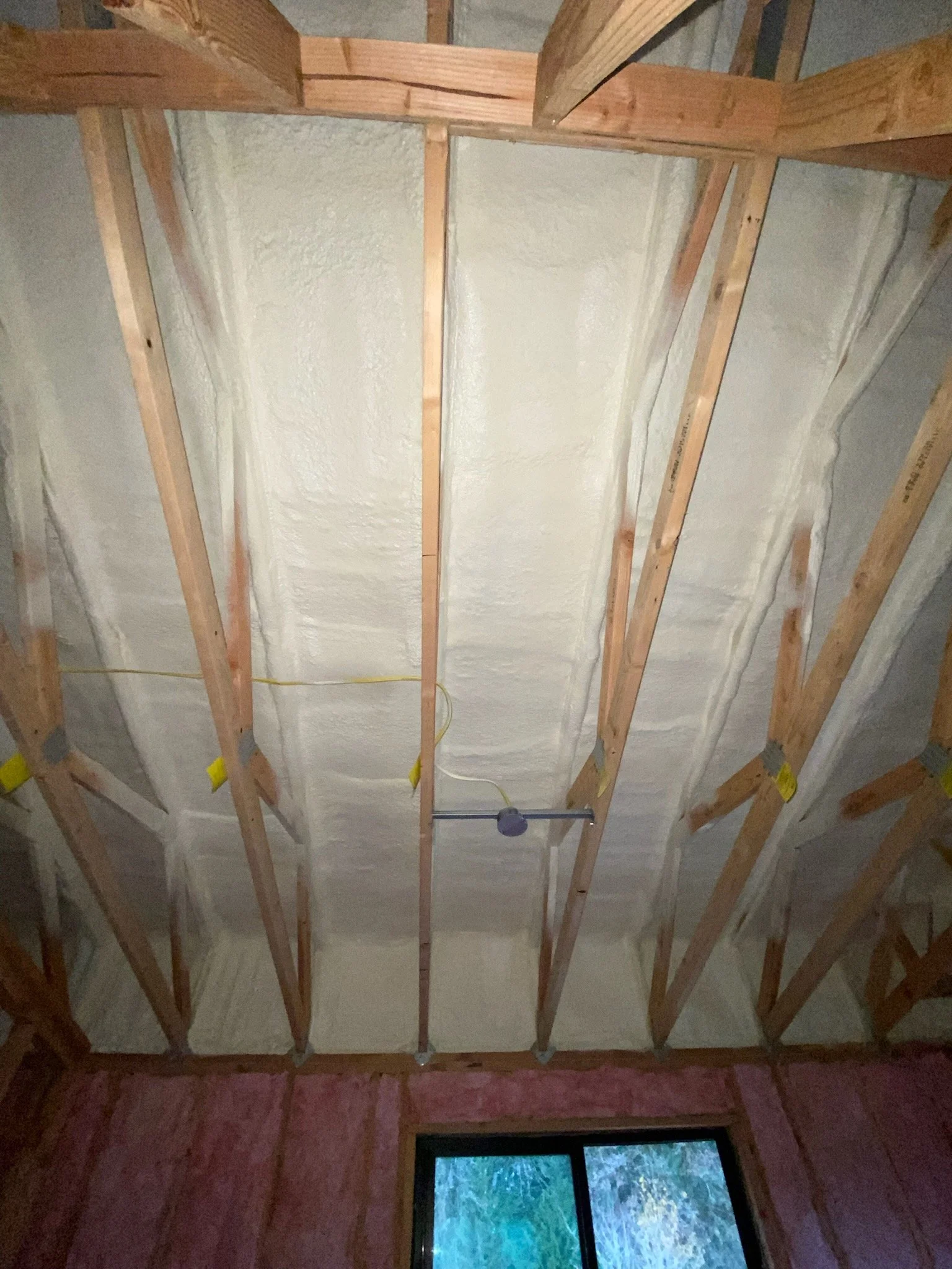 Construction of an attic ceiling with wooden beams and spray foam insulation, with a window visible below.