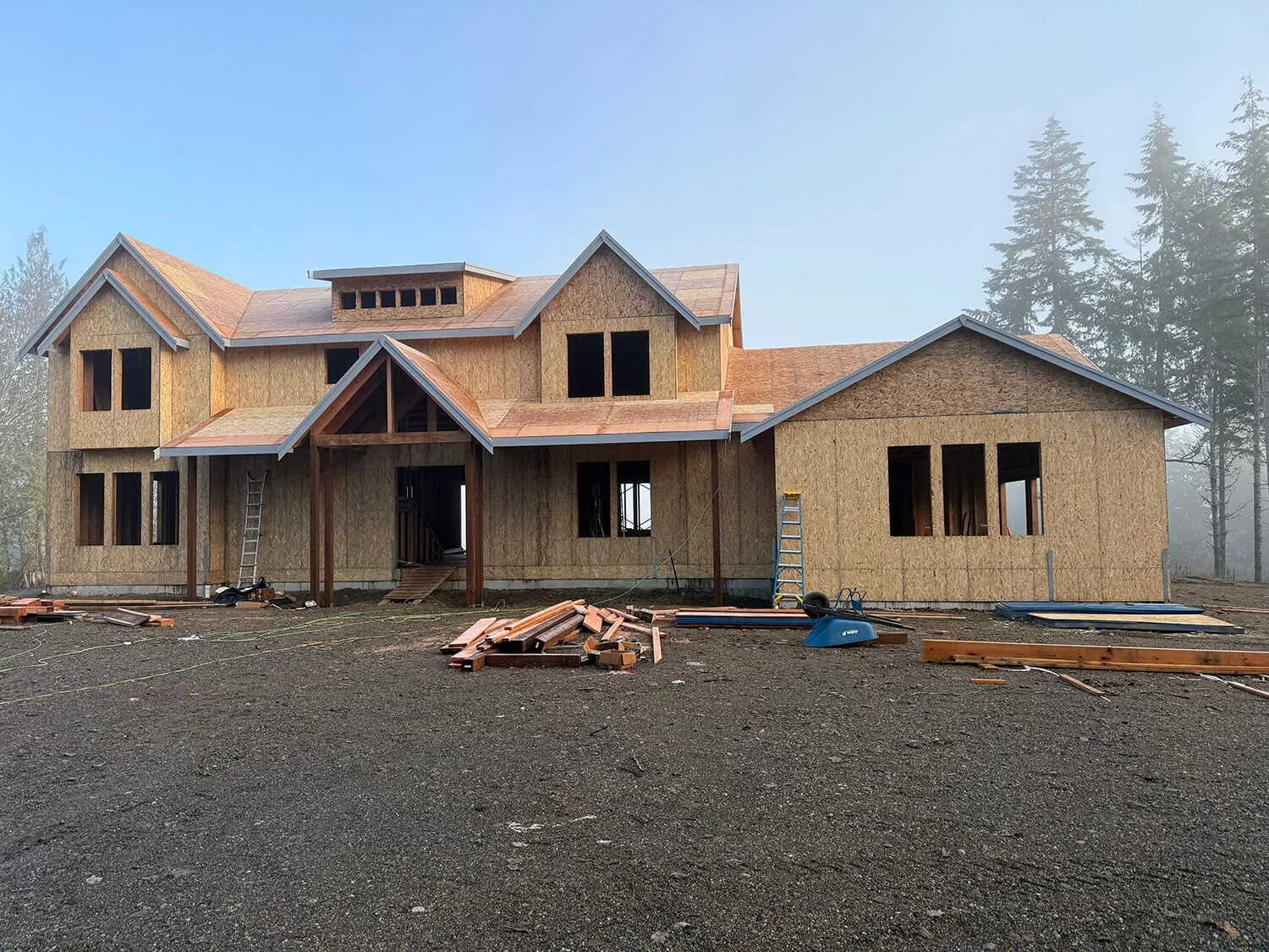 A house under construction with unfinished wooden exterior walls and roof framing, surrounded by construction debris and tools, with trees in the background.