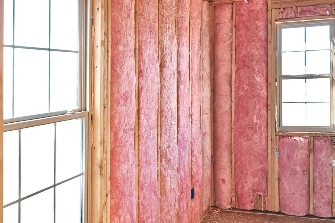 Interior of a room under construction with pink insulation in the walls, two windows, and exposed wooden studs.