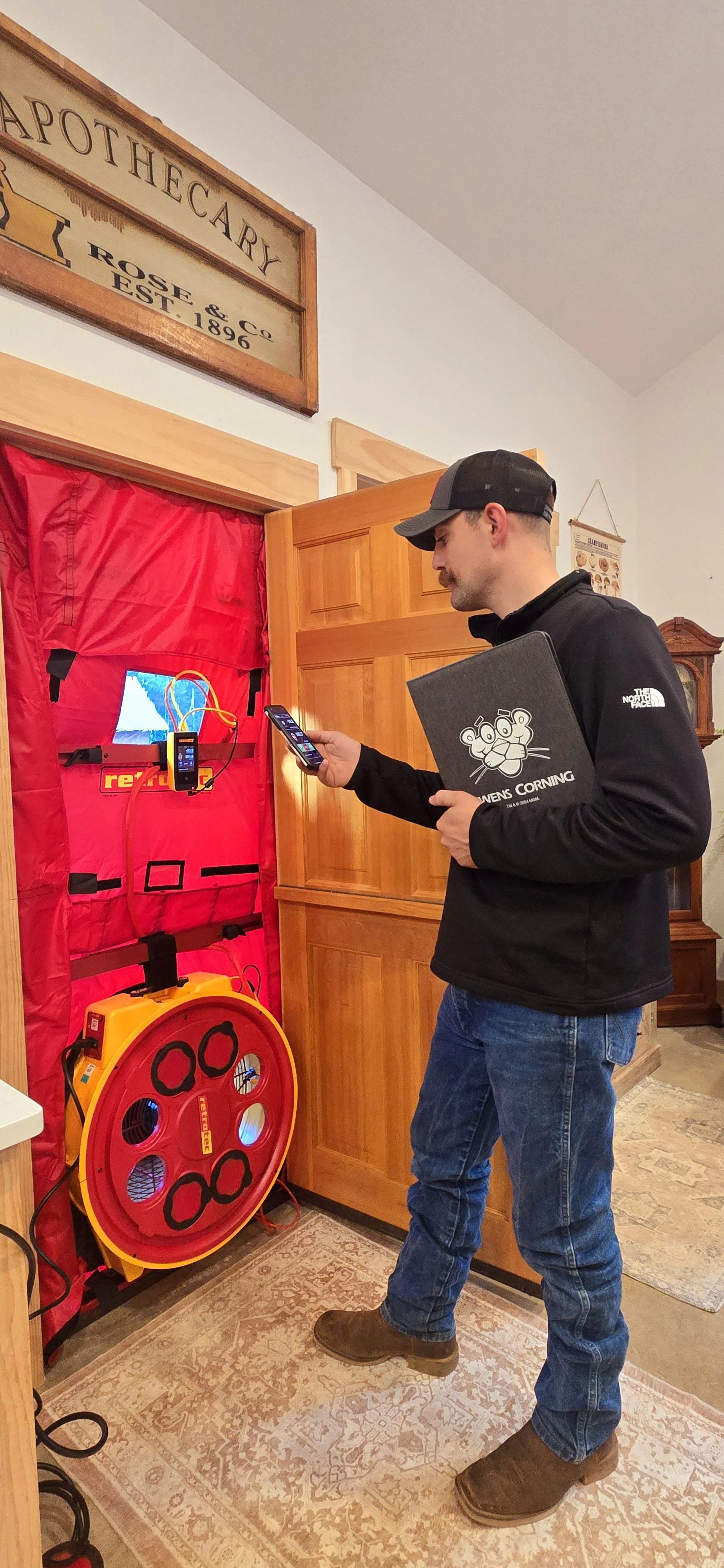 A man in casual clothing and a cap standing inside a room, using a smartphone with a speaker-like device nearby. The speaker is yellow and red and is connected to a small monitor and other electronic equipment inside a red padded enclosure. The room has wooden cabinets, a patterned rug, and a sign on the wall that reads 'Apothecary.'
