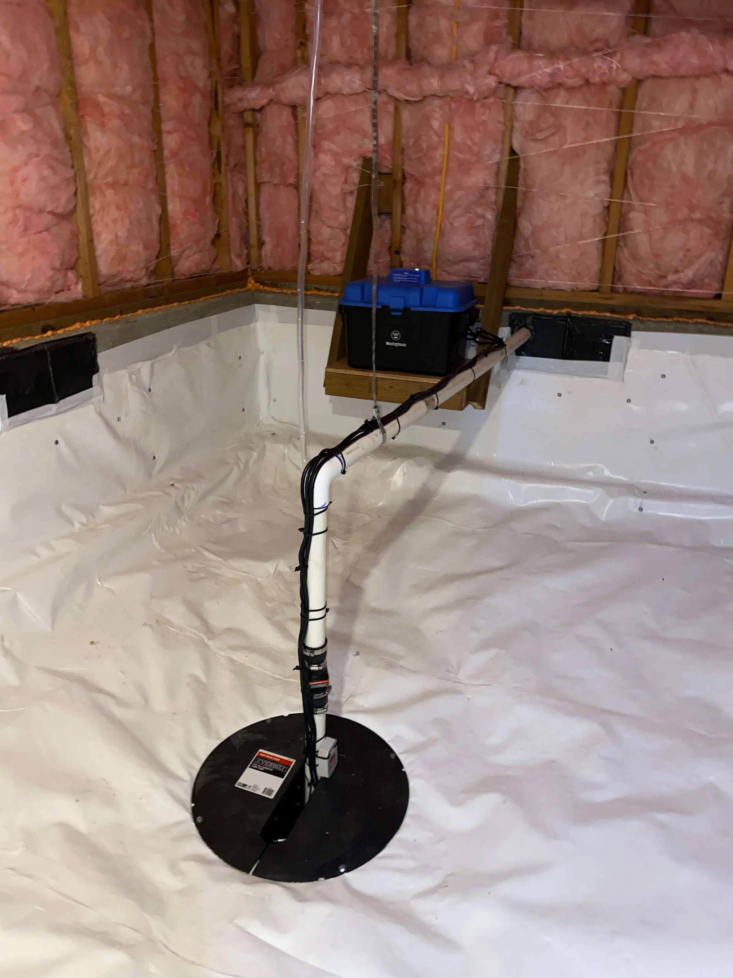 A residential basement crawl space with white vapor barrier on the floor, a sump pump and pink insulation