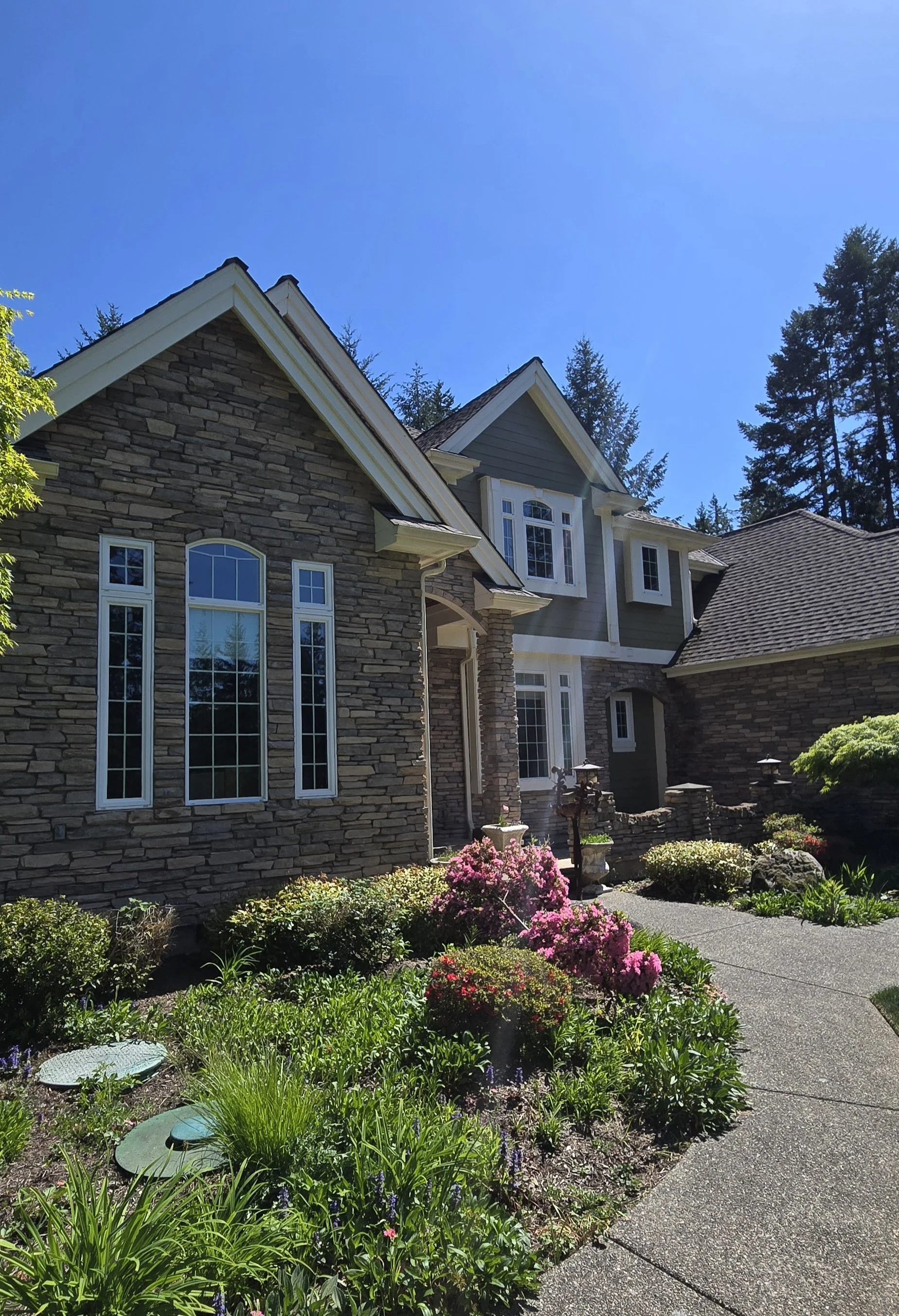 A house with stone and gray siding exterior, large decorative windows, and a lush garden with colorful flowers and shrubs, under a clear blue sky.