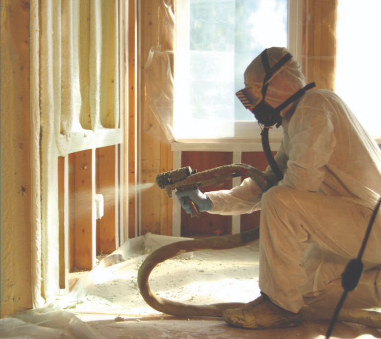 A professional spray foam technician in full protective gear, including a suit, gloves, and a mask installing closed-cell spray foam onto the interior wall of a residential home under construction.