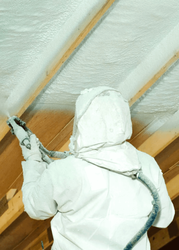 Person in protective gear installing spray foam insulation into a ceiling cavity from below.