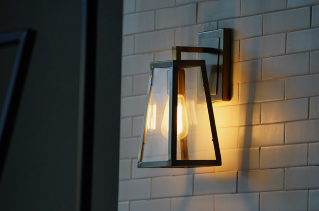 Wall-mounted modern lantern-style light fixture with an exposed bulb on a white tiled wall.