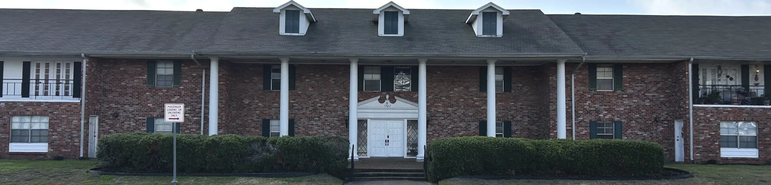 A two-story brick apartment building with white columns supporting a porch roof, black shutters, and dormer windows on the roof. There is a set of steps leading to the main entrance, surrounded by shrubbery, and a