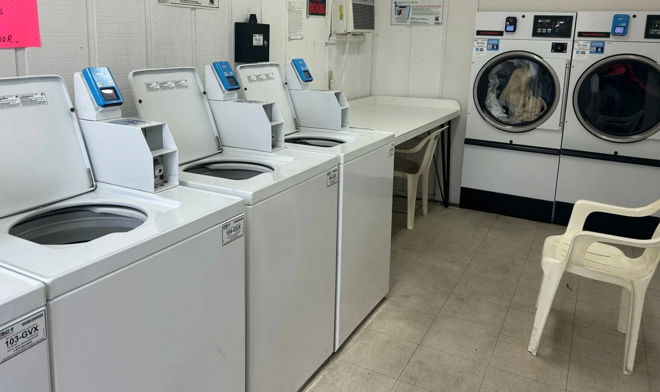 A laundromat with several top-loading washing machines, and two front-loading dryers. There is a white plastic chair and a small table in the corner.