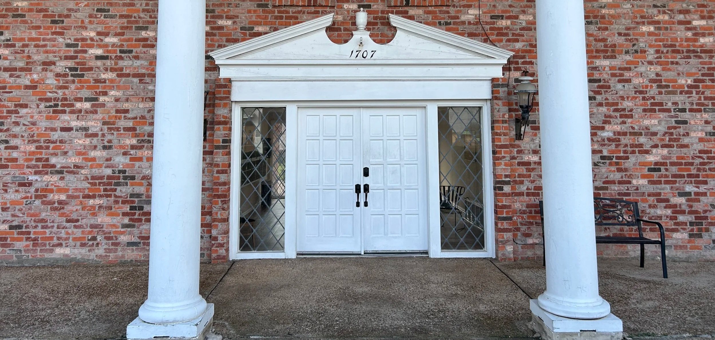 Entrance to a brick building with white double doors, white columns, and a triangular pediment with the year 1707 inscribed on it. There are glass windows on either side of the door and a bench on the right.