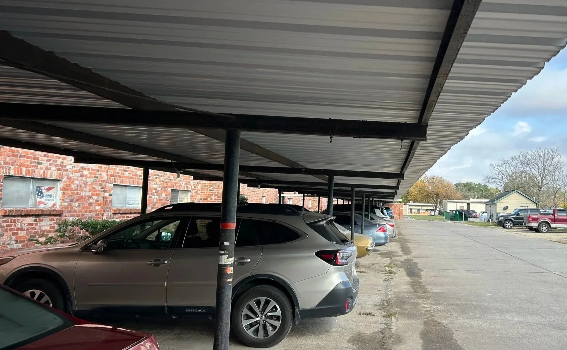 Parking lot with several parked cars under a metal awning, brick building with windows on the left, trees and houses in the background.