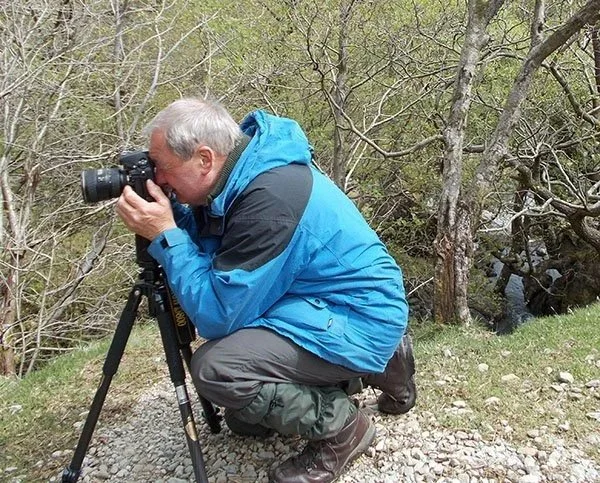 A man in outdoor gear using a camera on a tripod in a wooded area near a stream.