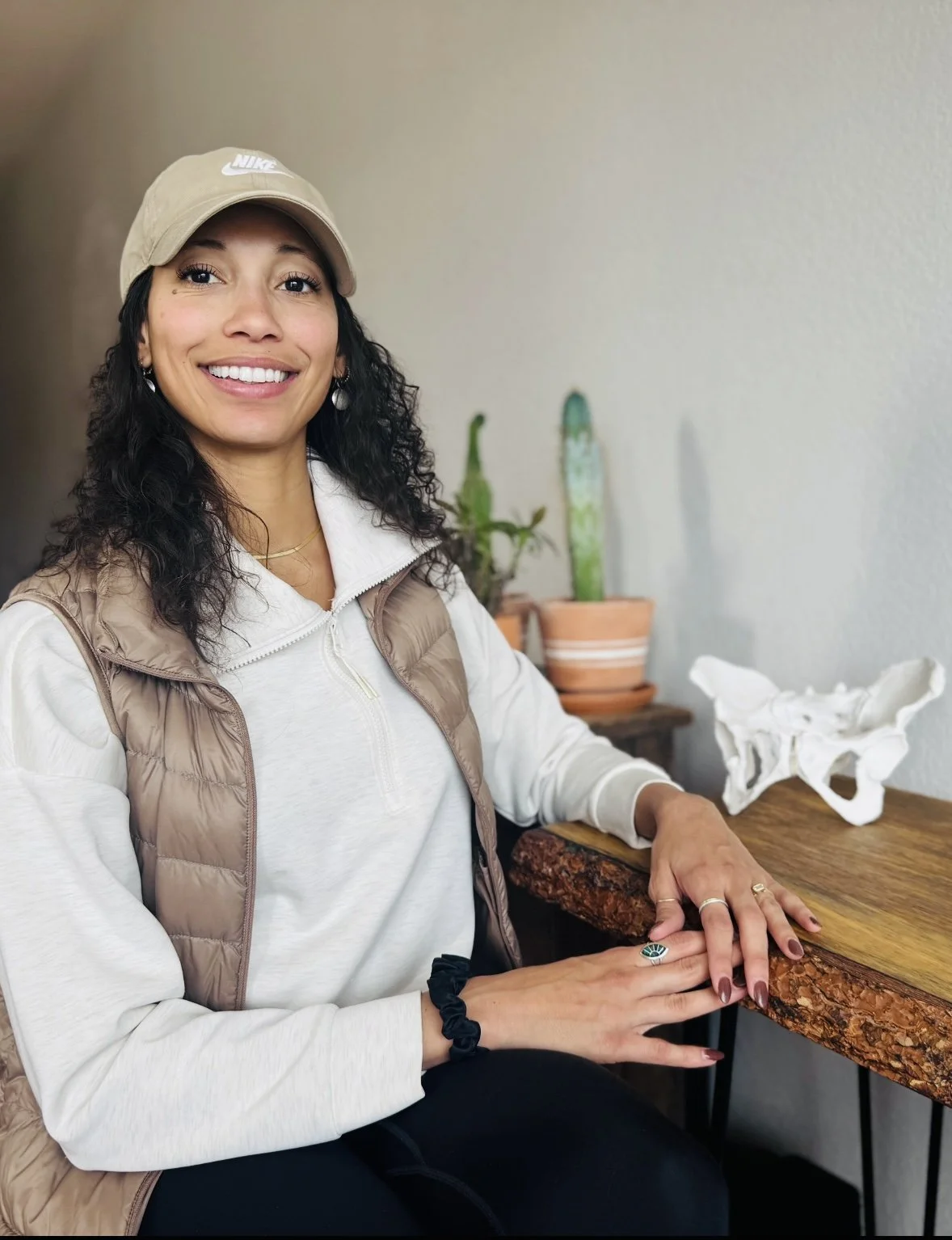 A woman with dark curly hair wearing a beige Nike cap, white jacket with brown vest, and jewelry, sitting at a wooden table with a smile.