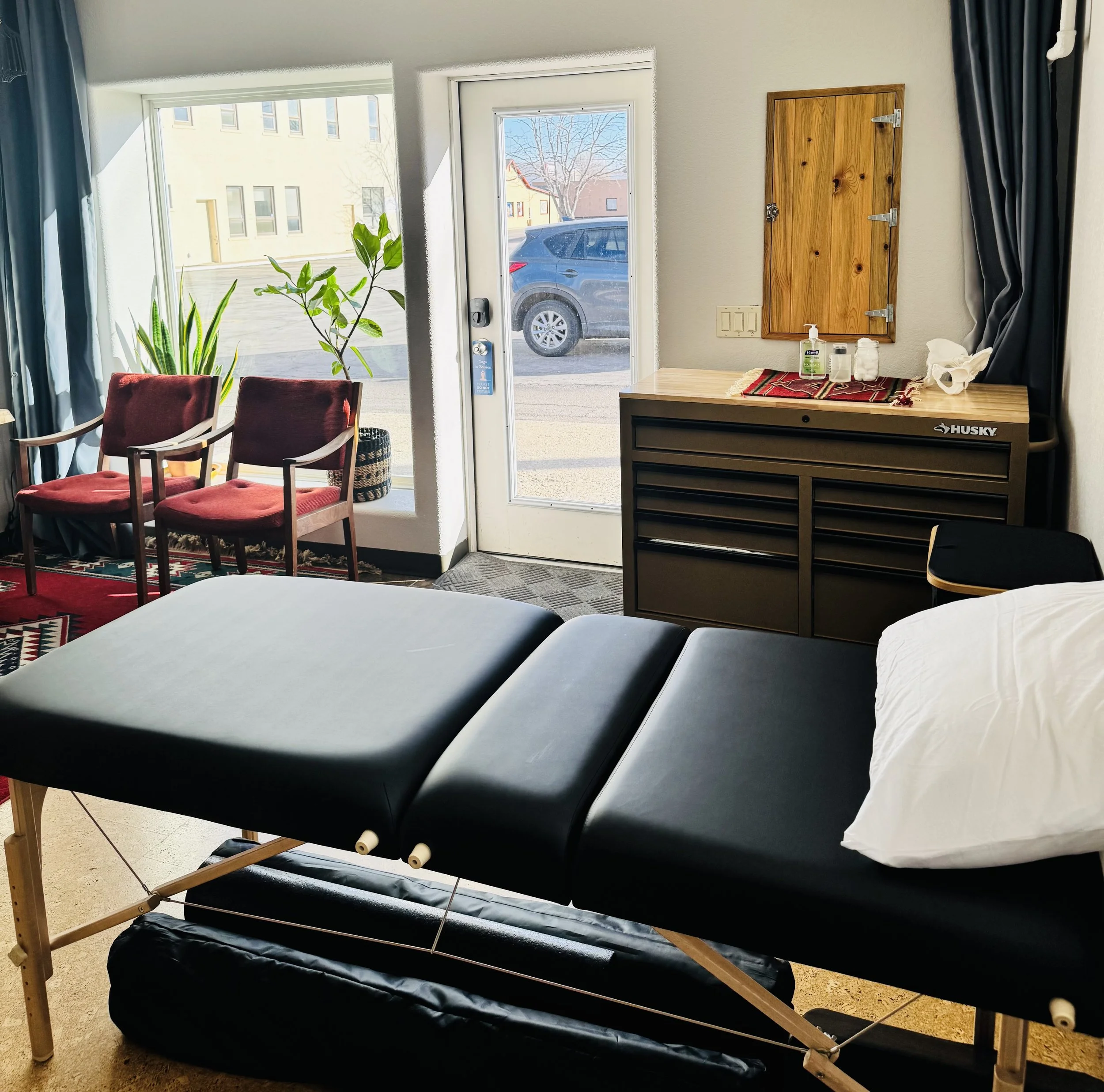 Interior of a therapy or massage room with a massage table covered in black fabric, a white pillow, a wooden cabinet with bottles and hand sanitizer, a small windowed door, two red chairs near a window with potted plants, and curtains.