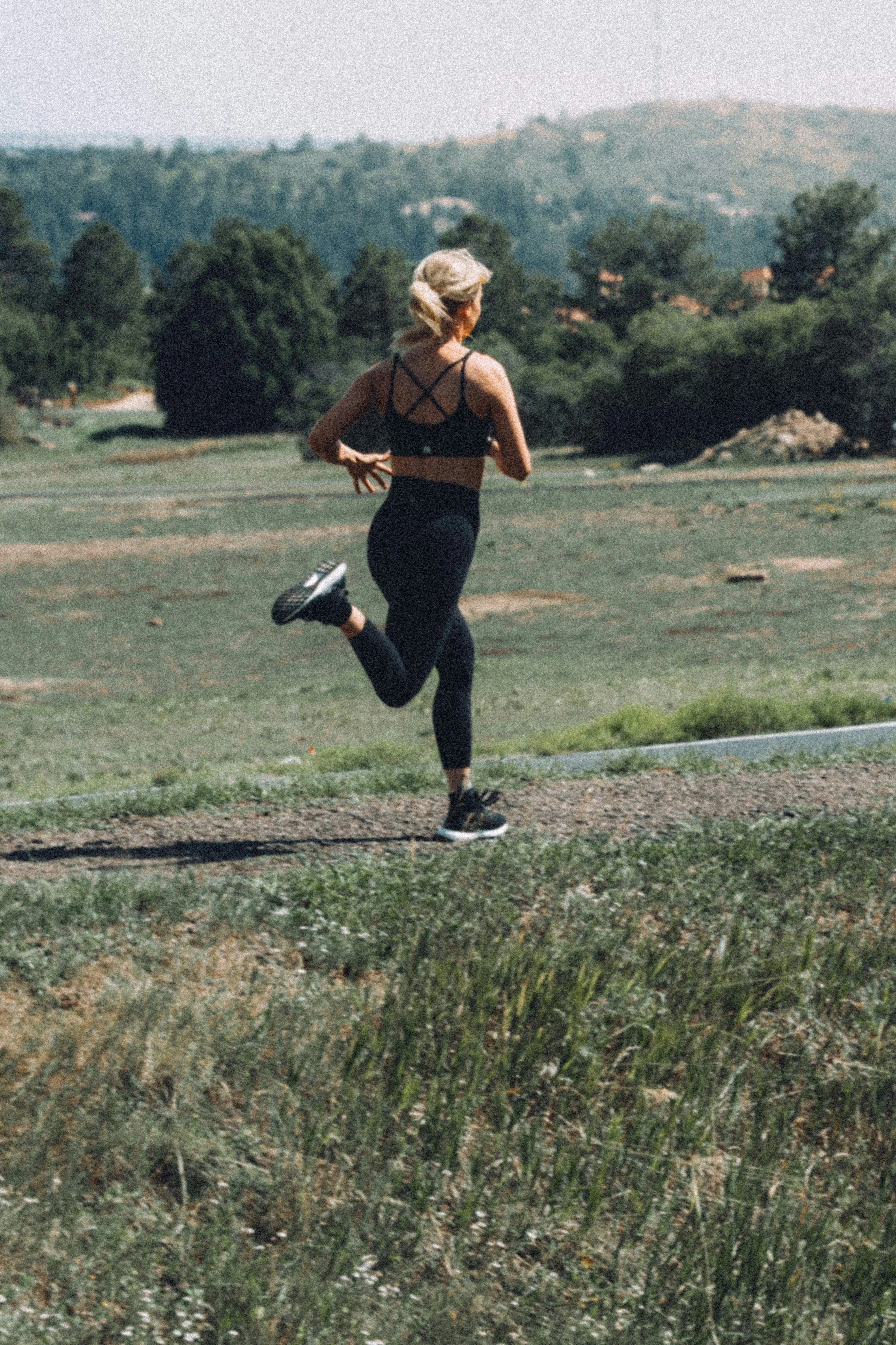 A woman running outdoors on a grassy trail in a rural area with green trees and hills in the background.