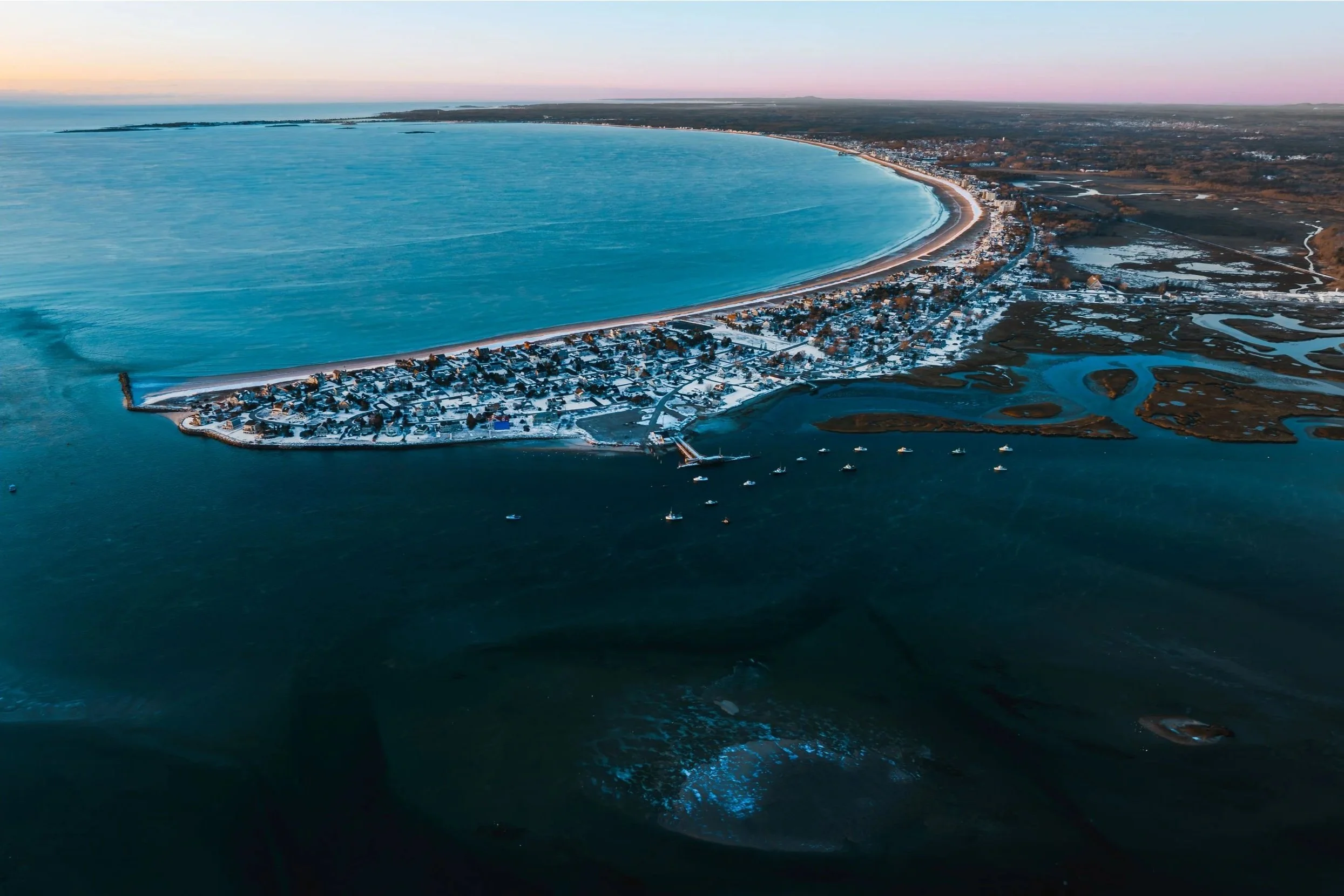 Aerial view of a snow-covered coastal town with a curved shoreline, boats anchored in the water, and wetlands or marshes to the right, under a pastel-colored sky at sunset.