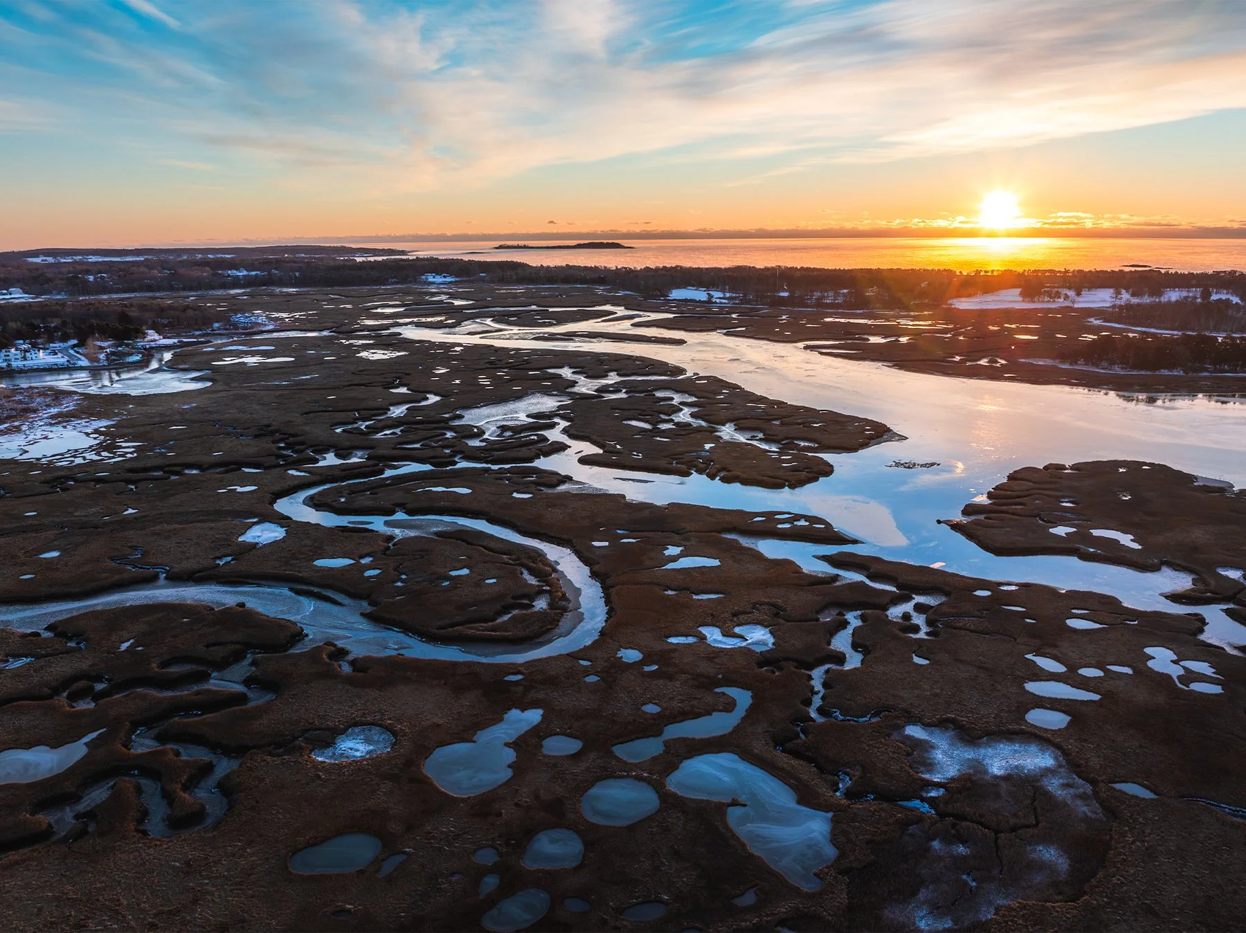 Aerial view of a river delta at sunset with winding waterways, patches of ice, and a sunset over the horizon.