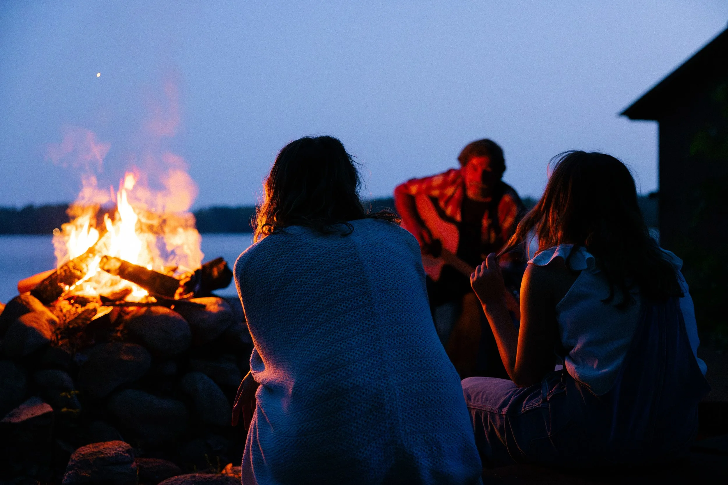 Three people sitting by a campfire near a lake at dusk, with one person playing guitar.