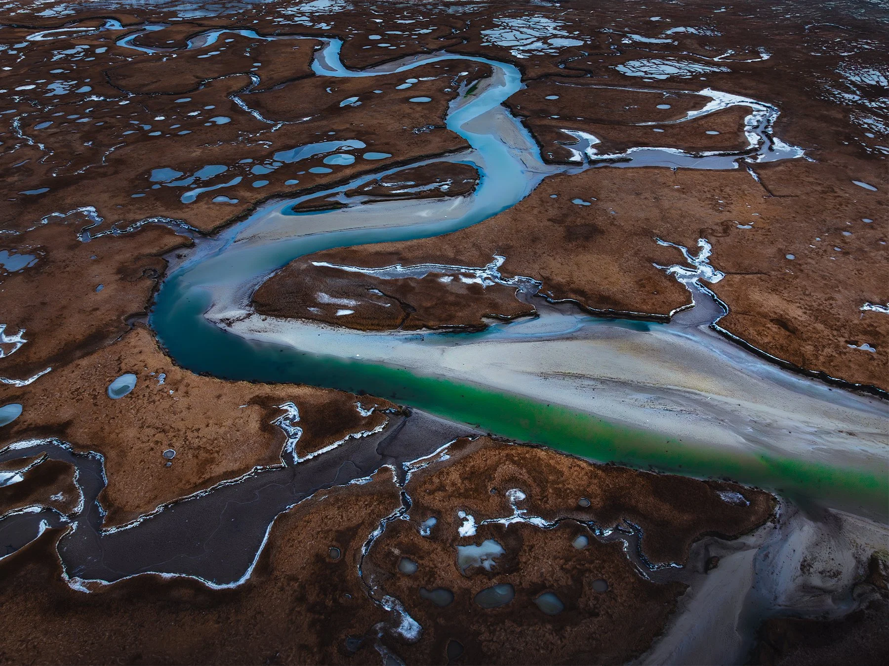 Aerial view of a winding river with blue and green hues flowing through a landscape of brown and white patches, possibly during winter or early spring.