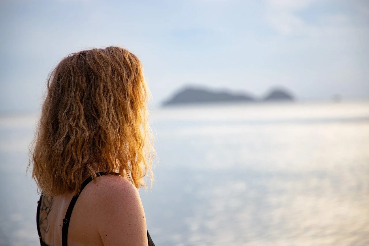 A woman with curly red hair looking out at the ocean during sunset or sunrise.