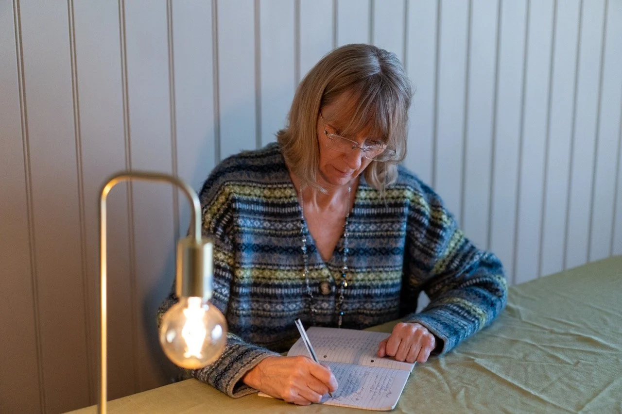 A woman with glasses writing in a notebook at a green table with a small lamp nearby.