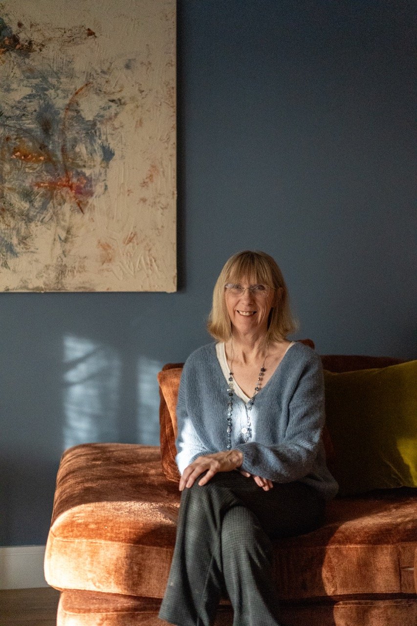 A smiling woman with glasses and shoulder-length blonde hair sitting on a brown velvet sofa in a room with blue walls, a colourful abstract painting on the wall, and sunlight creating shadows on the wall.