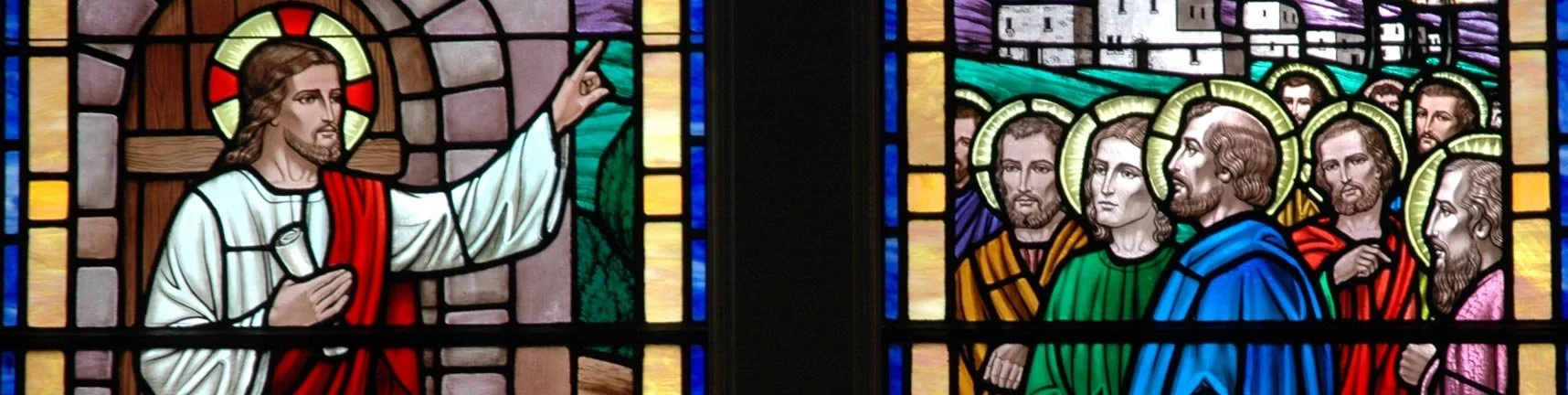 Stained glass window depicting Jesus Christ teaching, with Him pointing upwards, and a group of people listening, all with haloed heads.